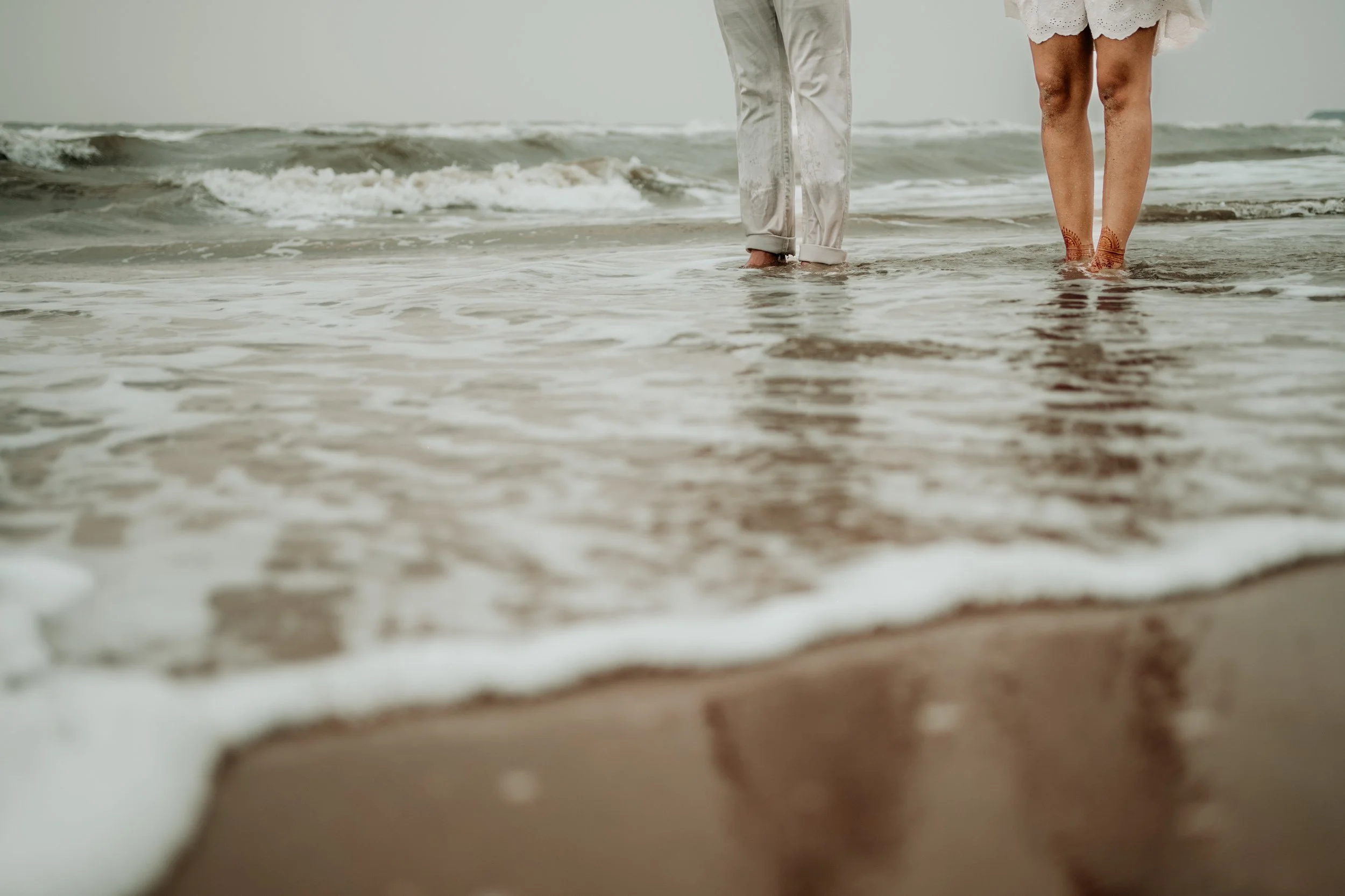 People standing in the shallow ocean water at the beach, with their legs and feet visible, wearing light-colored clothing.