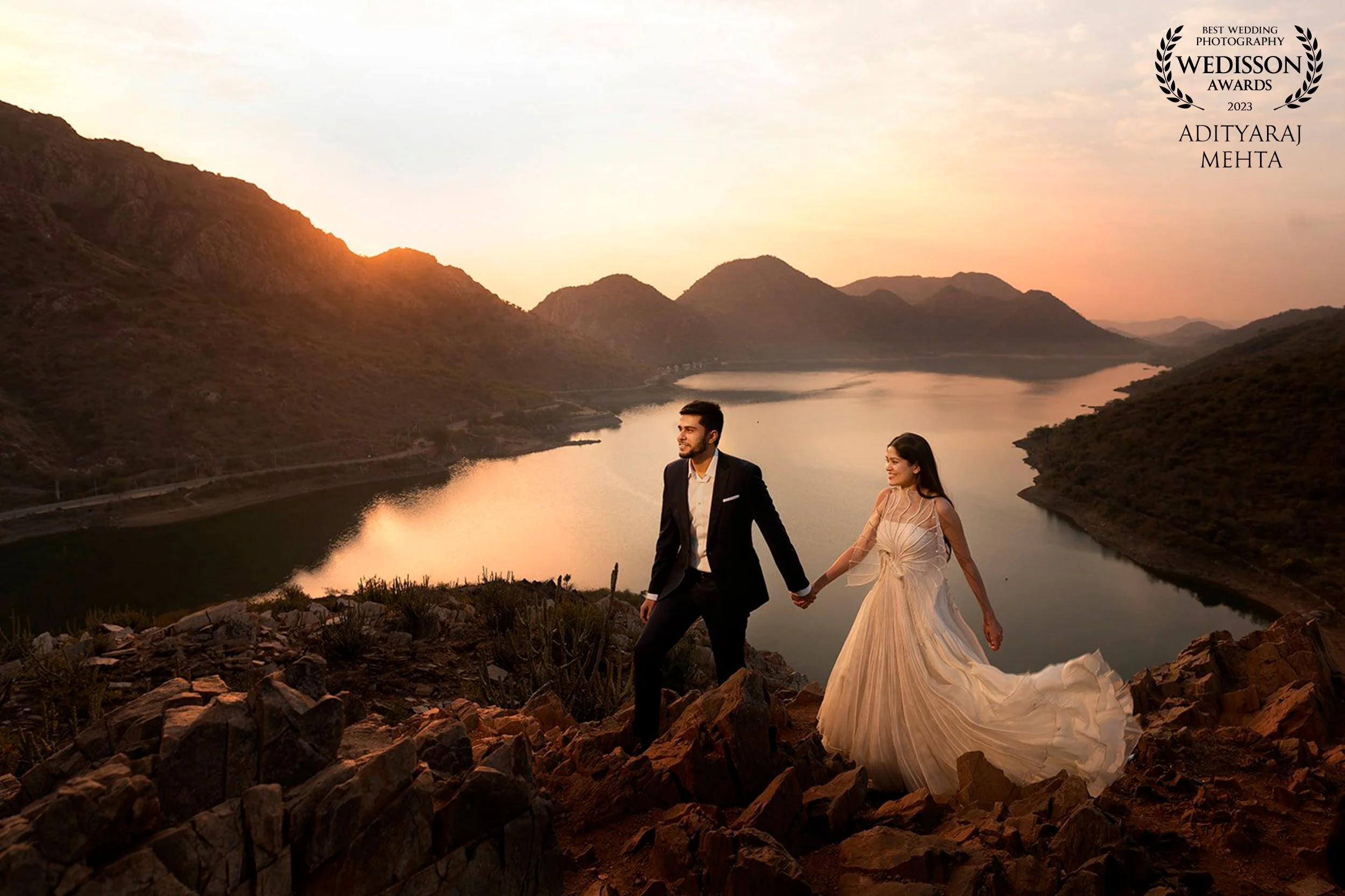 A newlywed couple holding hands on rocky terrain overlooking a river and mountains at sunset, with a wedding photography award badge in the top right corner.