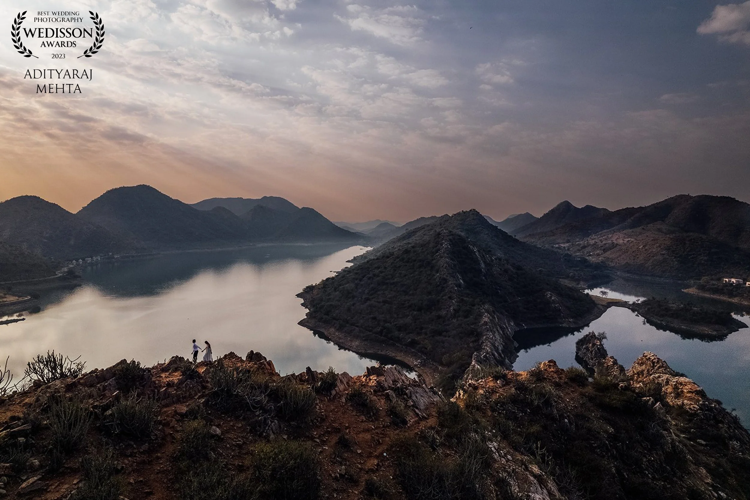 A panoramic view of a lake surrounded by mountains and hills during sunset or sunrise. Two people stand on the rocky foreground, holding hands. The sky is partly cloudy with warm hues near the horizon.