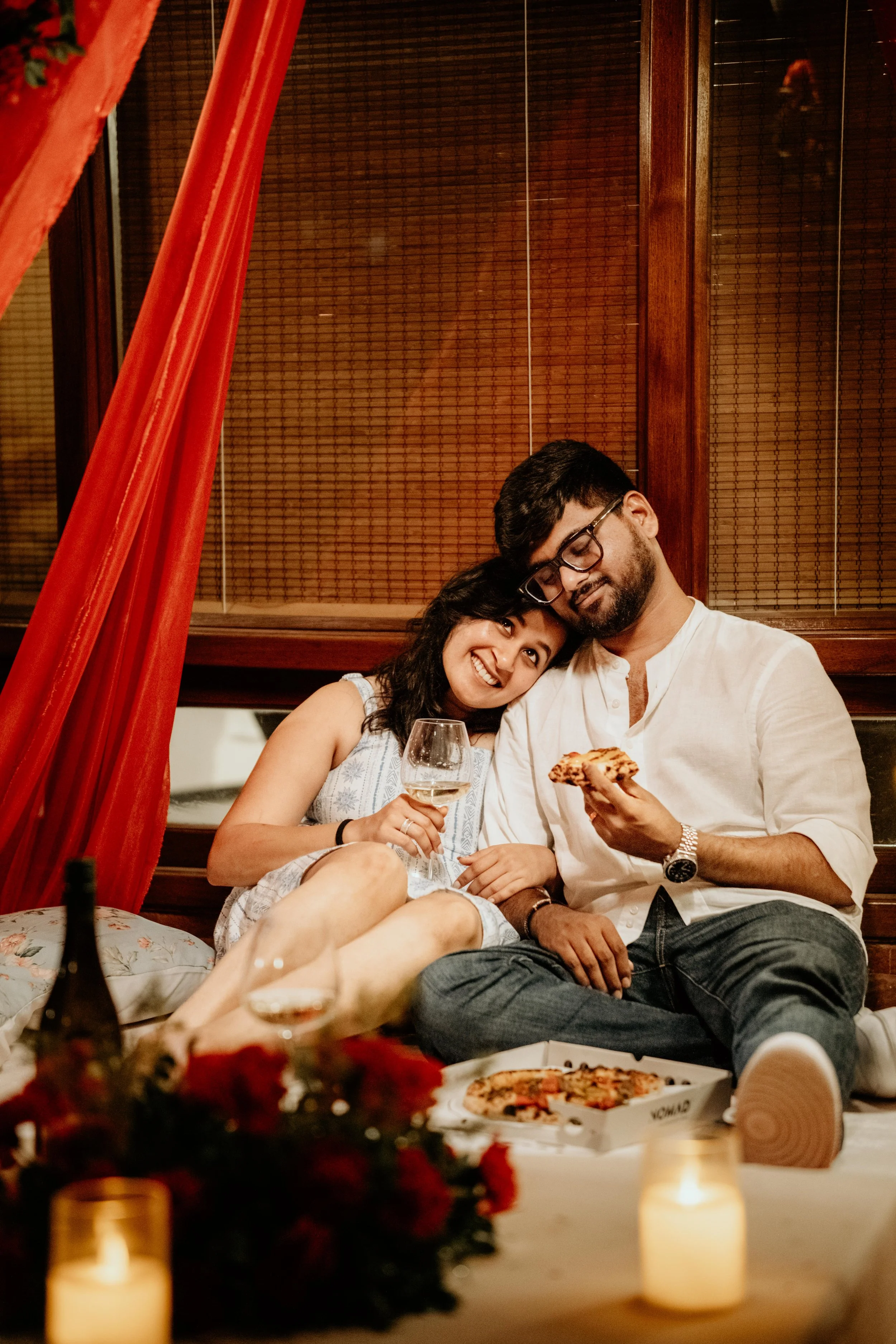 A couple sitting on a couch with pizza and wine, smiling and relaxing at home with candles and a bouquet of flowers in the foreground.