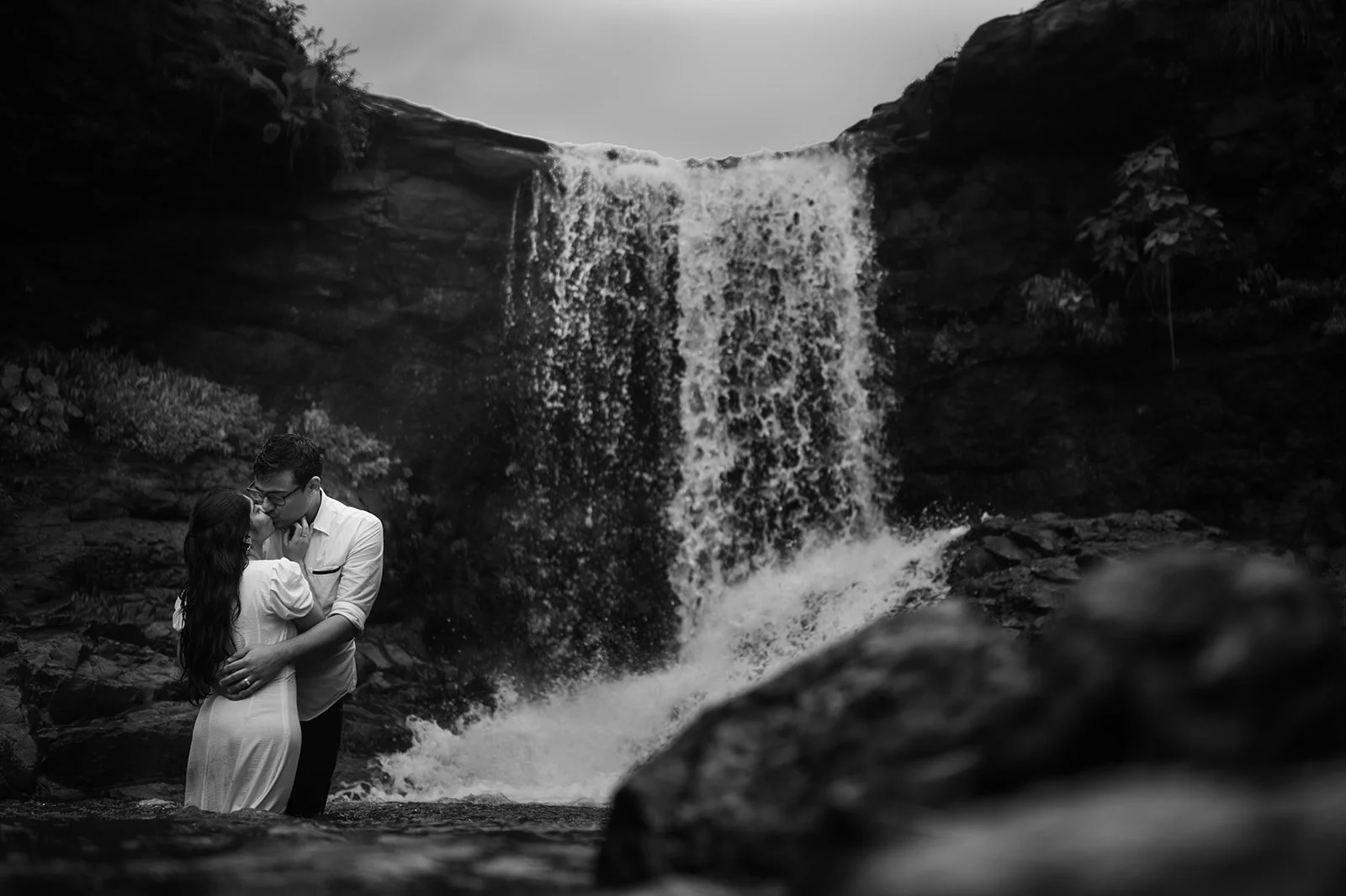 A couple embracing and about to kiss in front of a waterfall in a black and white photograph
