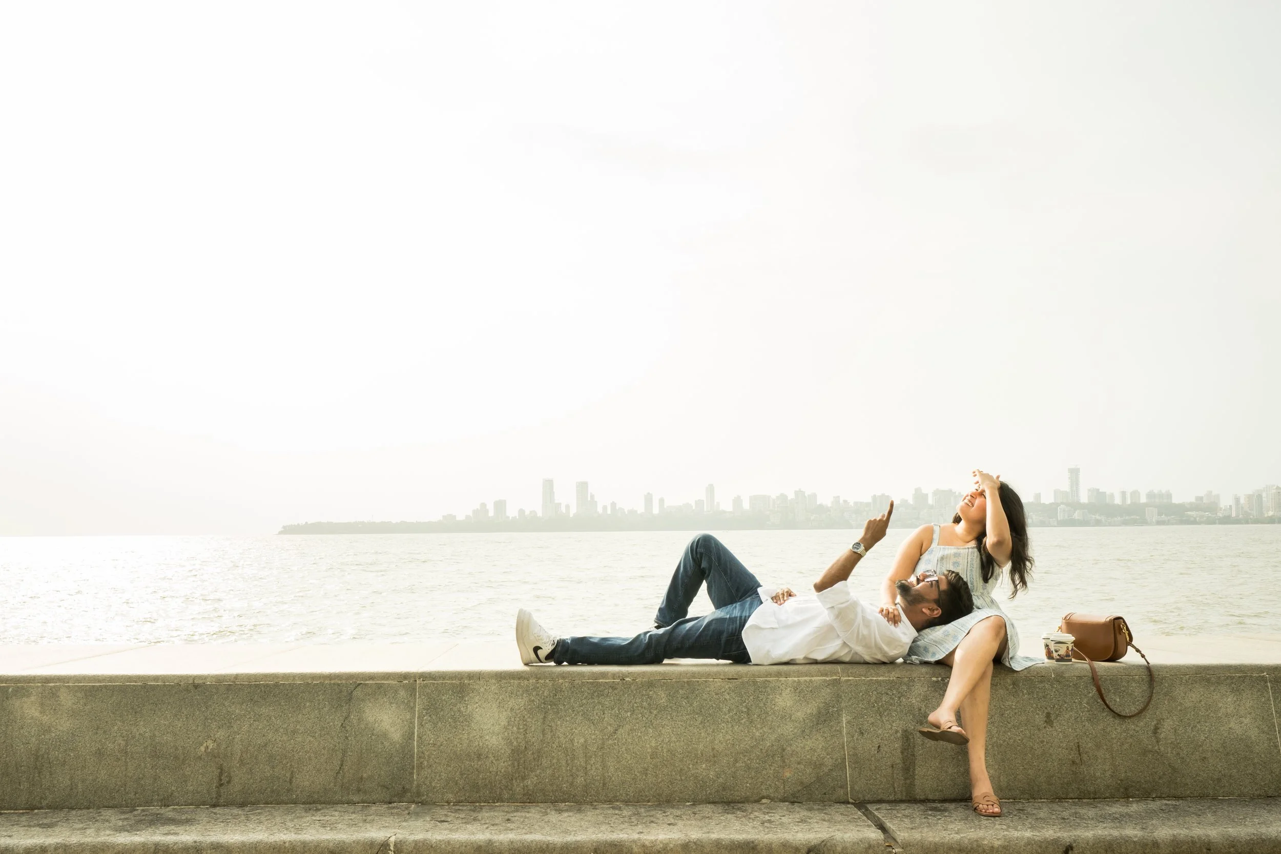 A man and woman relaxing on a concrete ledge by the water with a city skyline in the background, sharing a joyful moment.