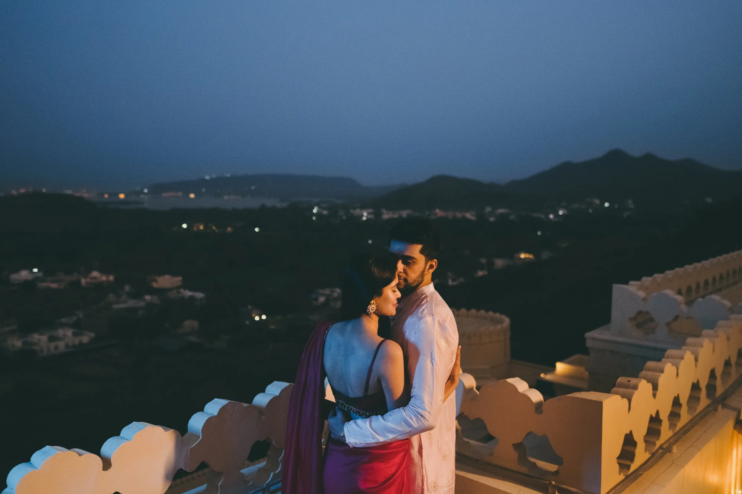 A couple dressed in traditional Indian attire embraces on a balcony overlooking a scenic landscape at dusk.