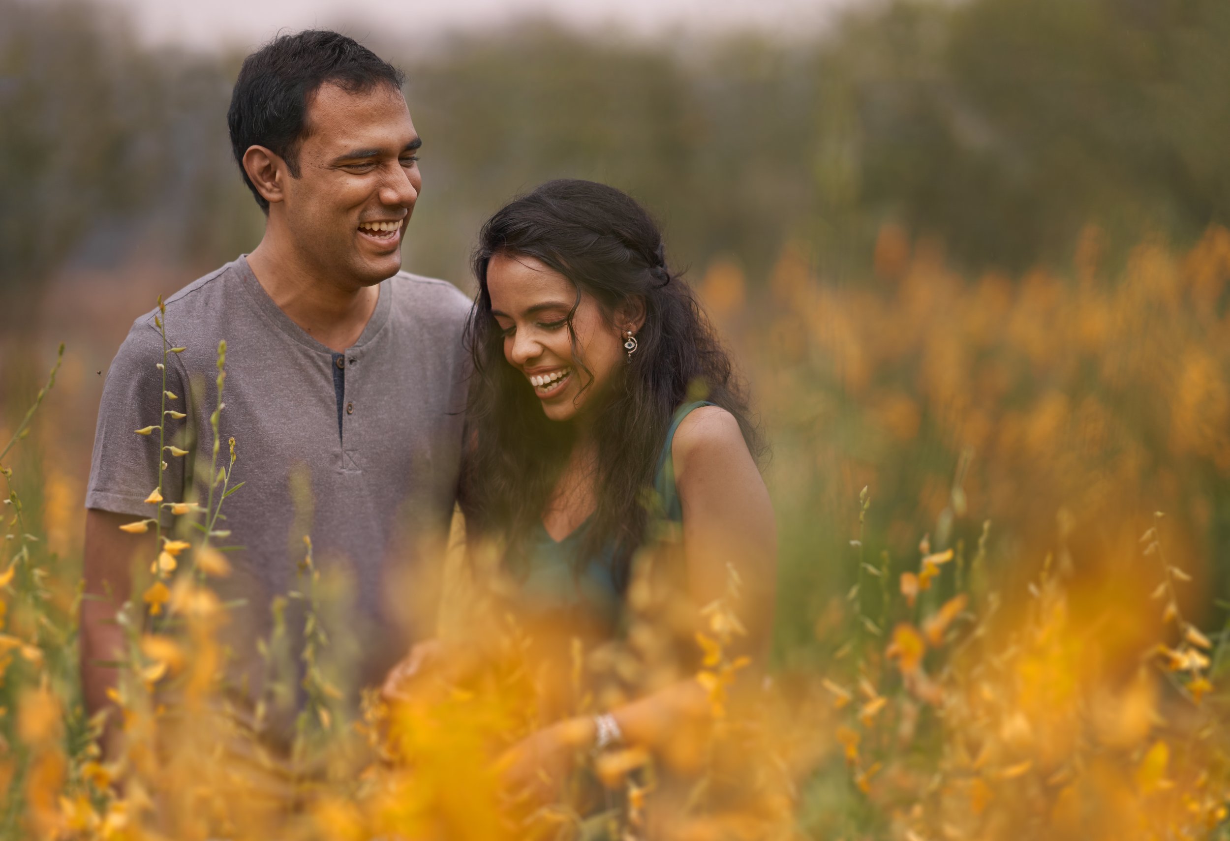A man and woman smiling and laughing together in a field of yellow flowers during daytime.