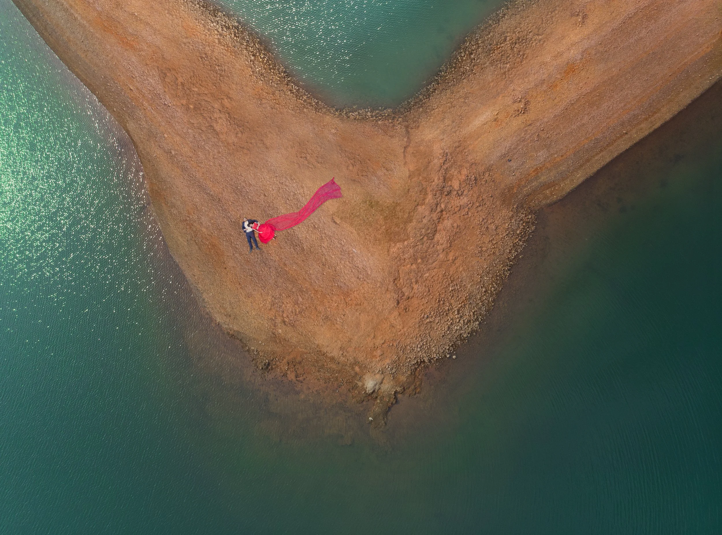 An aerial view of a couple lying on a small, curved, sandy landmass surrounded by water. The woman is wearing a long red dress, and the man is wearing a dark shirt and pants.