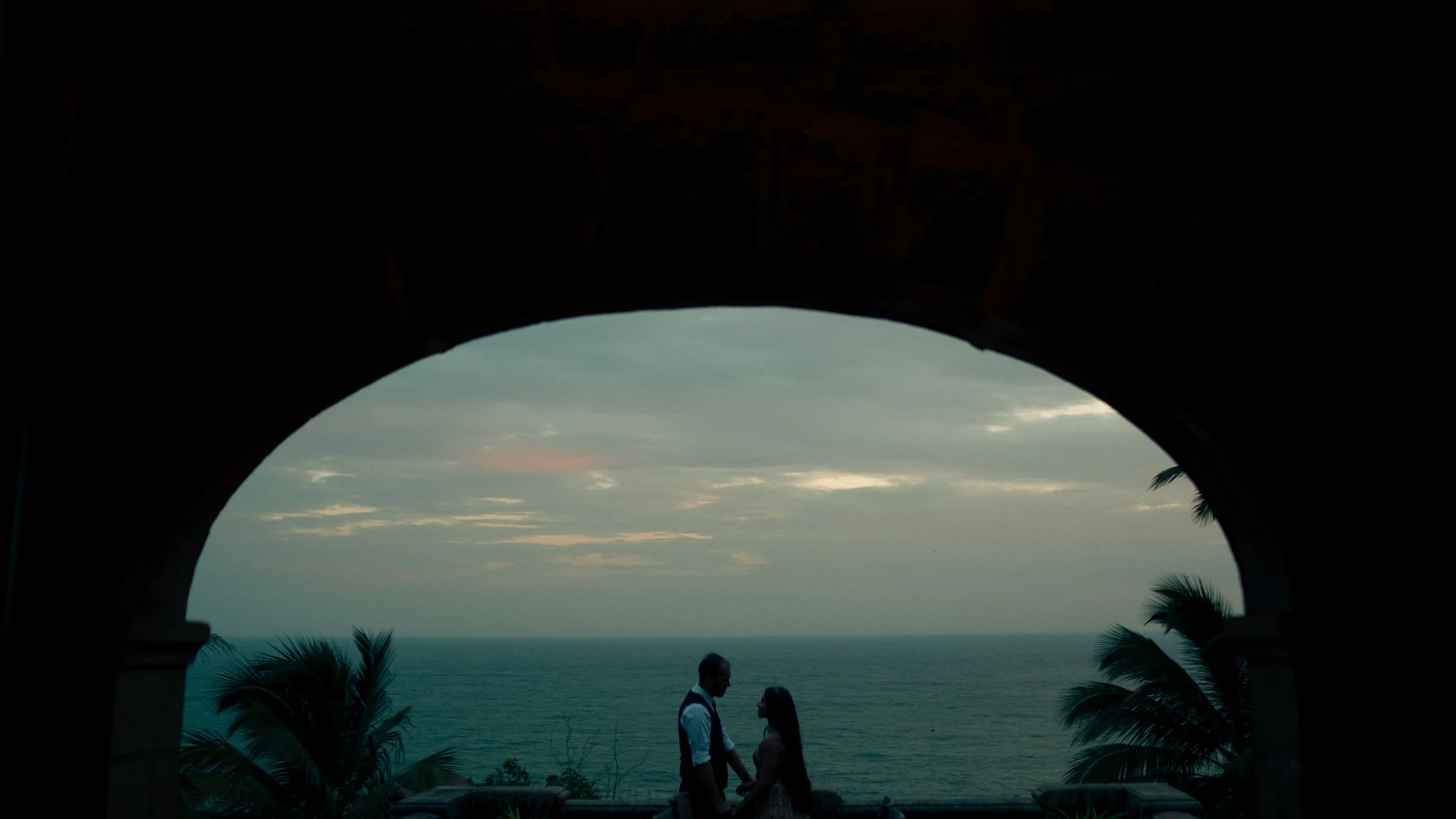 A couple holding hands and looking at each other on a balcony with a view of the ocean at sunset, framed by a stone arch and palm trees.