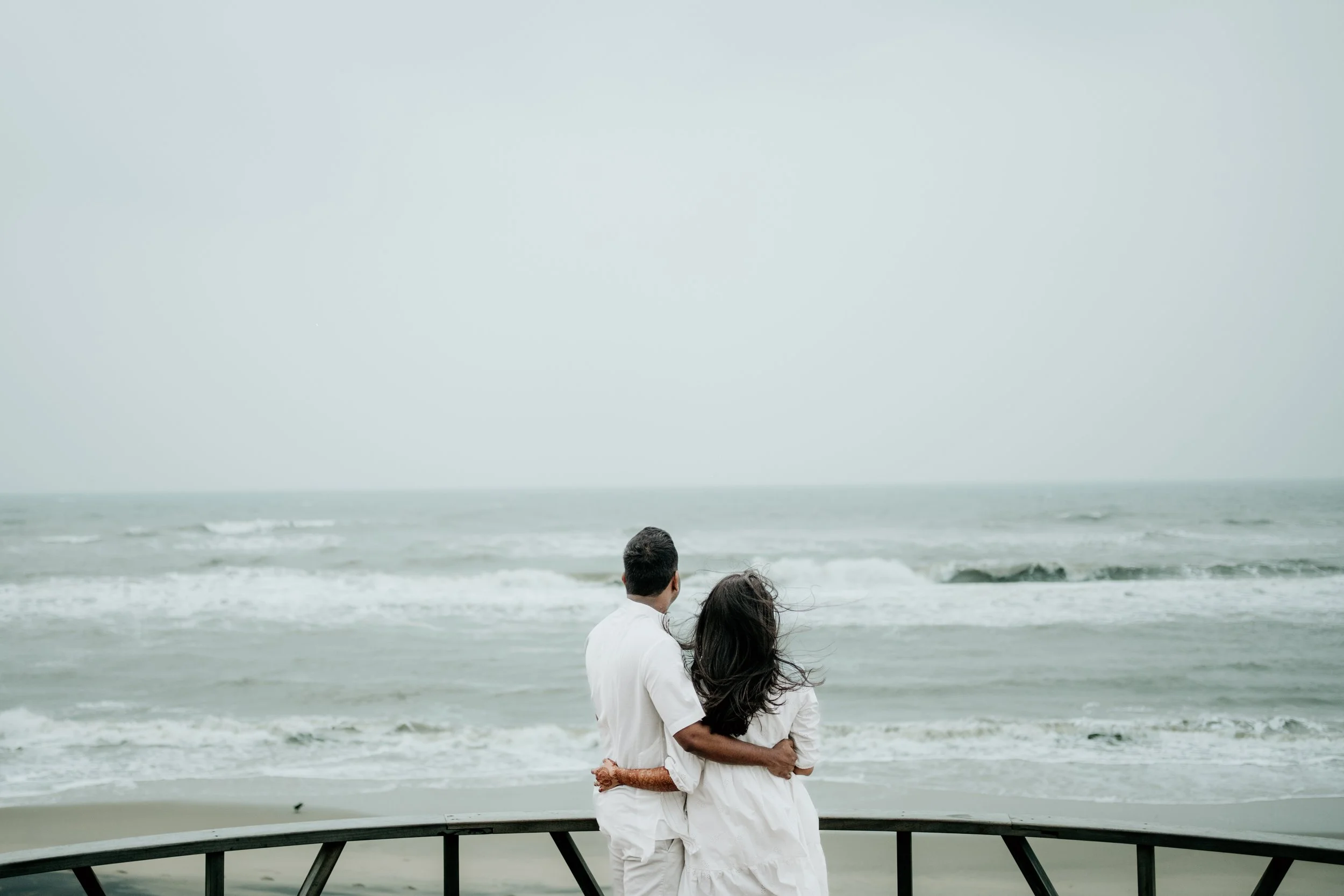 A couple stands close together on a beach, looking out at the ocean, with a cloudy sky overhead.