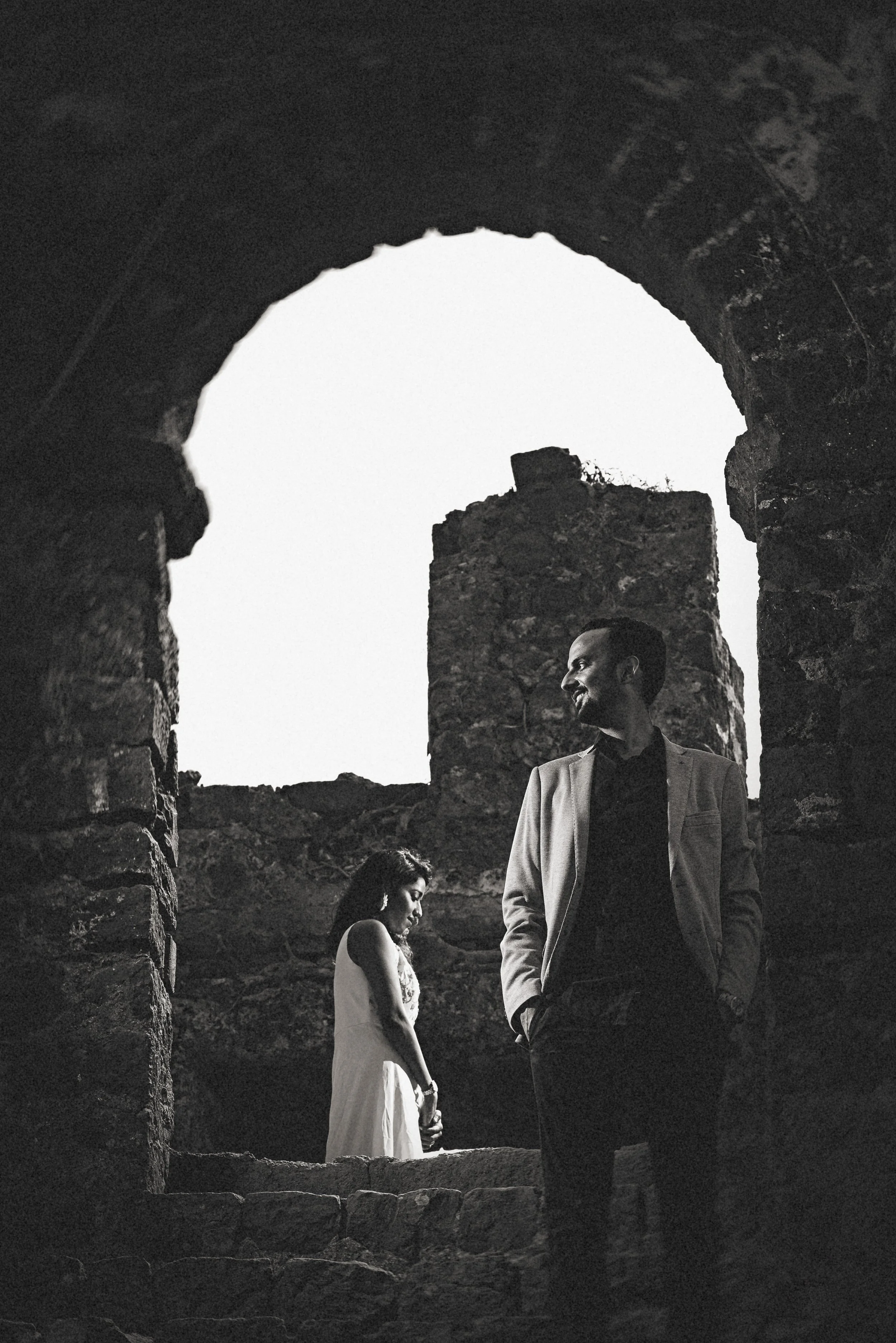 Black and white photo of a man and woman standing in a ruined stone structure, with the man smiling and looking off to the side while the woman looks down.