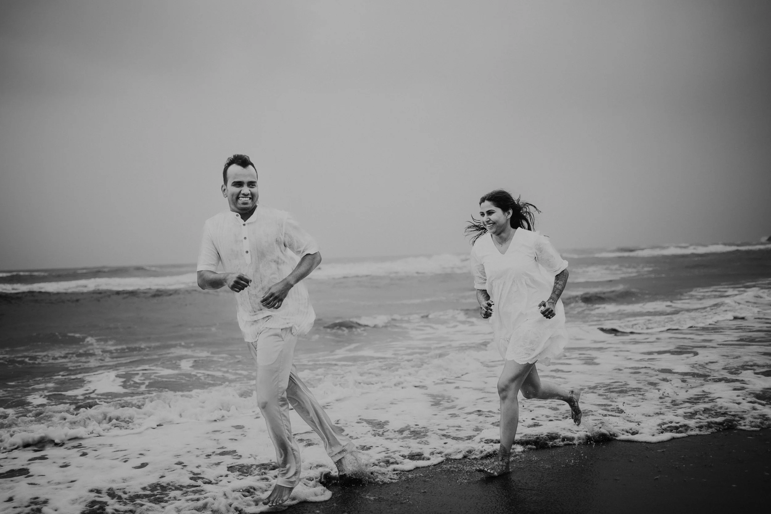 A couple running and smiling at the beach in black and white, near the shoreline with waves crashing around their feet.