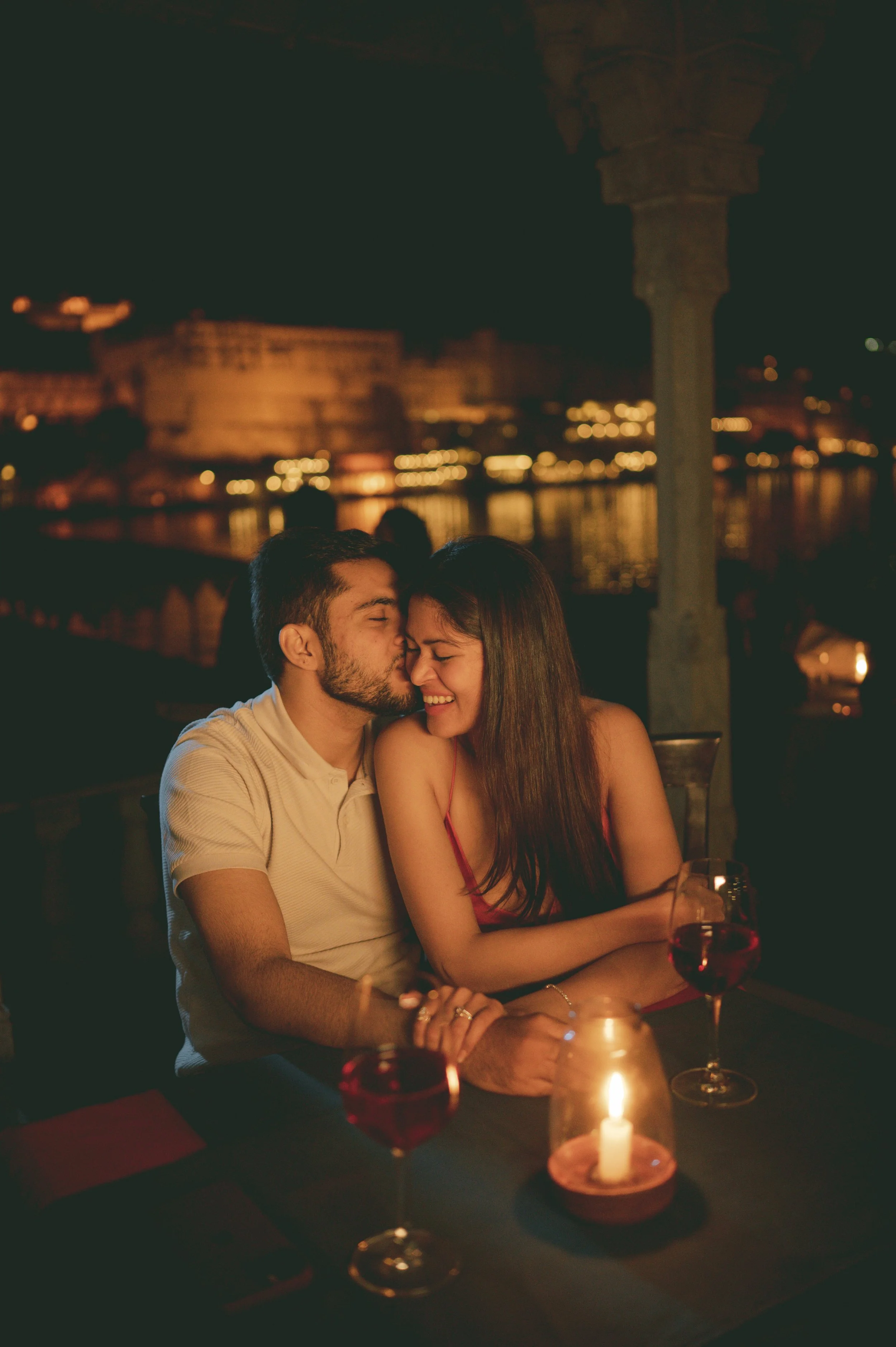 A romantic couple sitting at a candlelit dinner outdoors at night, with city lights and a body of water in the background.