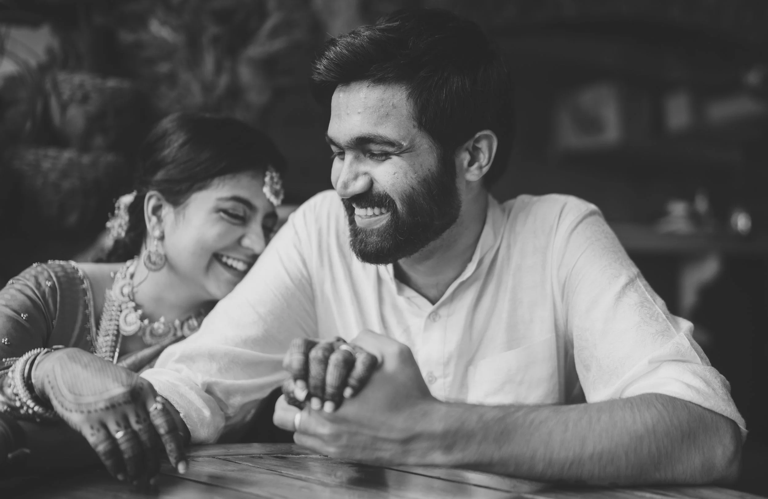 A man and woman with traditional Indian attire sharing a joyful moment, smiling and leaning close to each other, sitting at a table.
