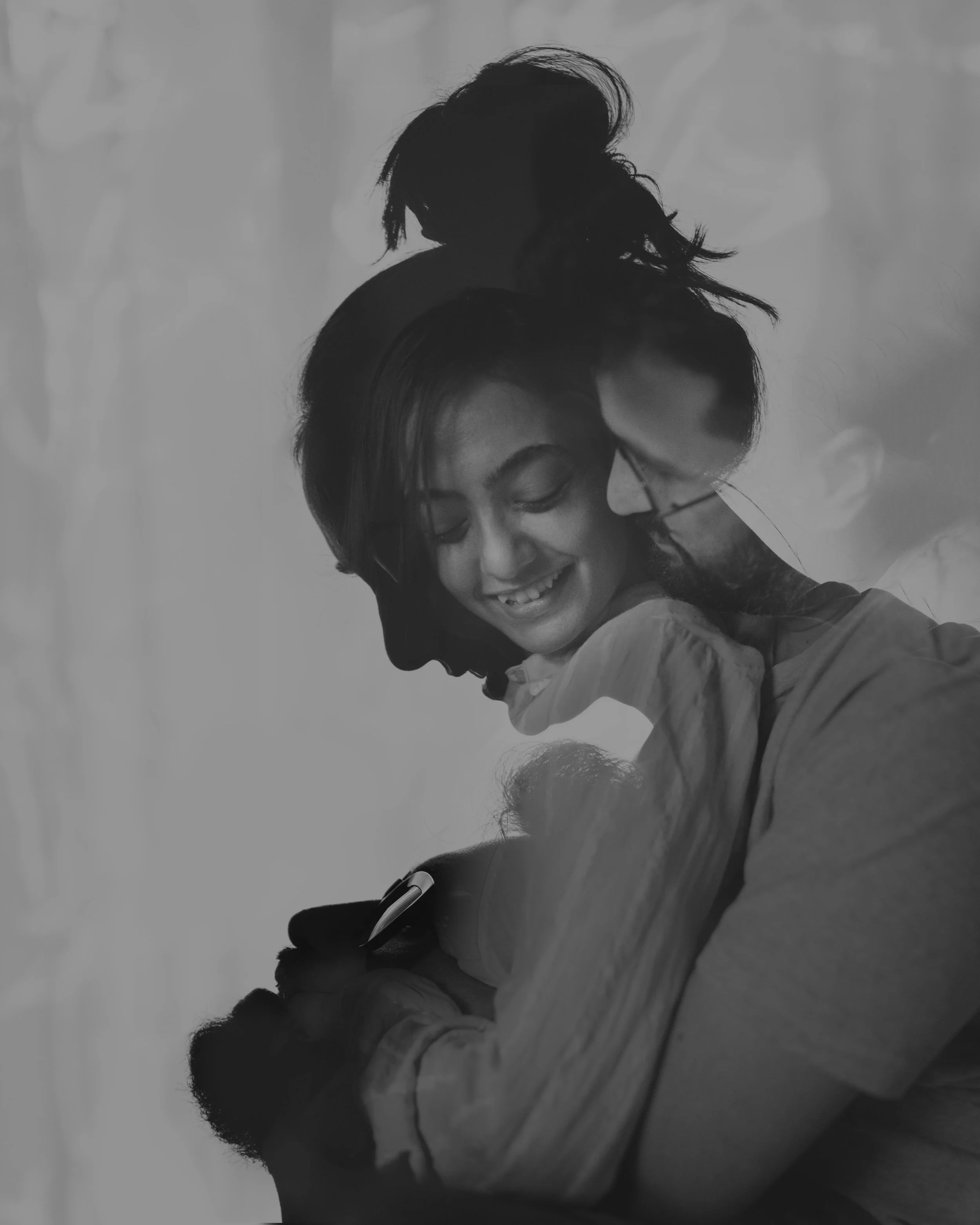 A black-and-white photo of a woman smiling and looking at her phone while a man leans over her shoulder, both appearing happy and affectionate.