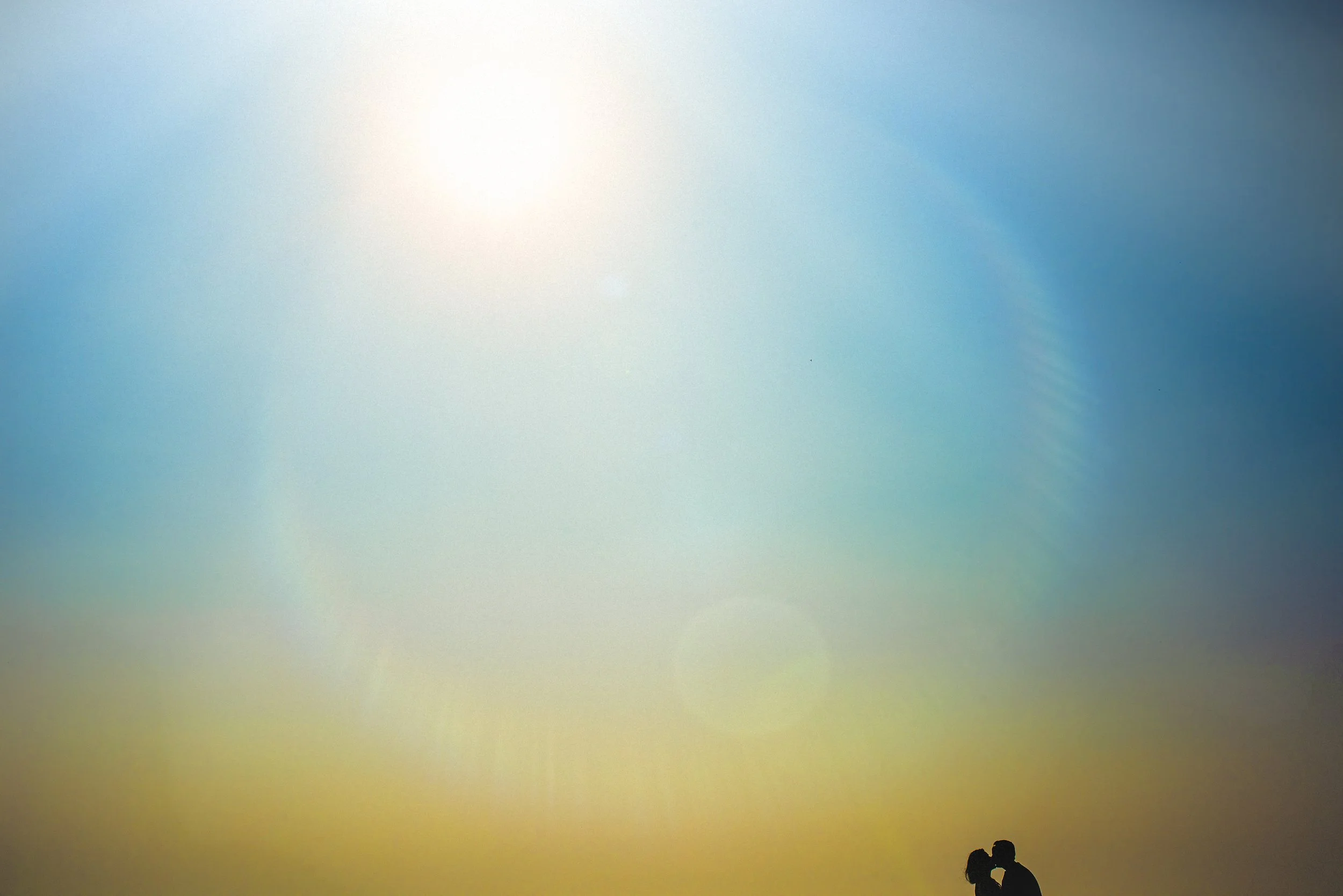 A silhouette of a couple kissing on the beach at sunset with a bright sun and soft rainbow lens flare in the sky.