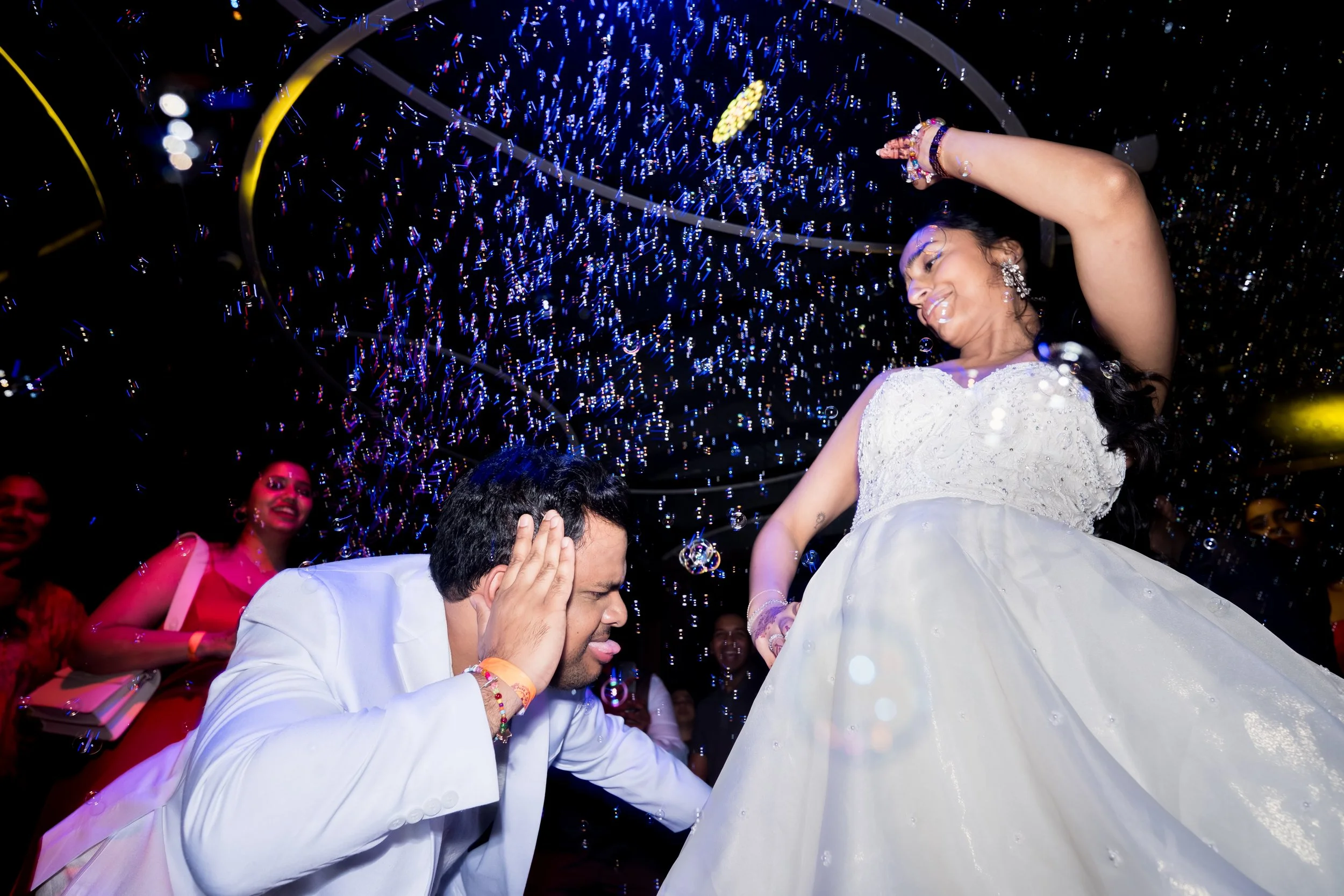 A woman in a white wedding dress dances with her hand raised, smiling, while a man in a white suit kneels before her with his hands on the sides of his head during a celebration with colorful lighting and confetti.