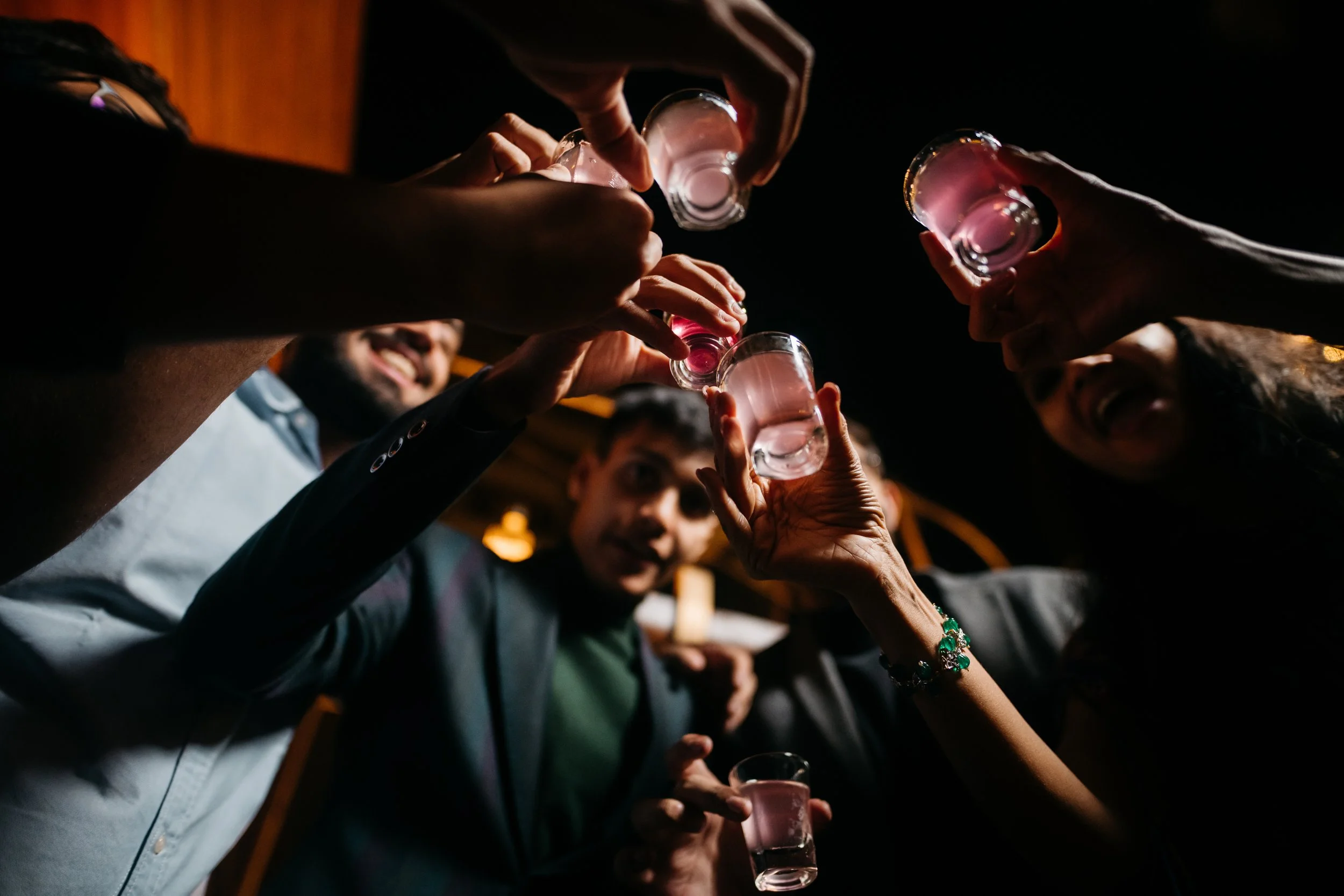 Group of friends toasting with shot glasses of pink drink in a dark indoor setting.