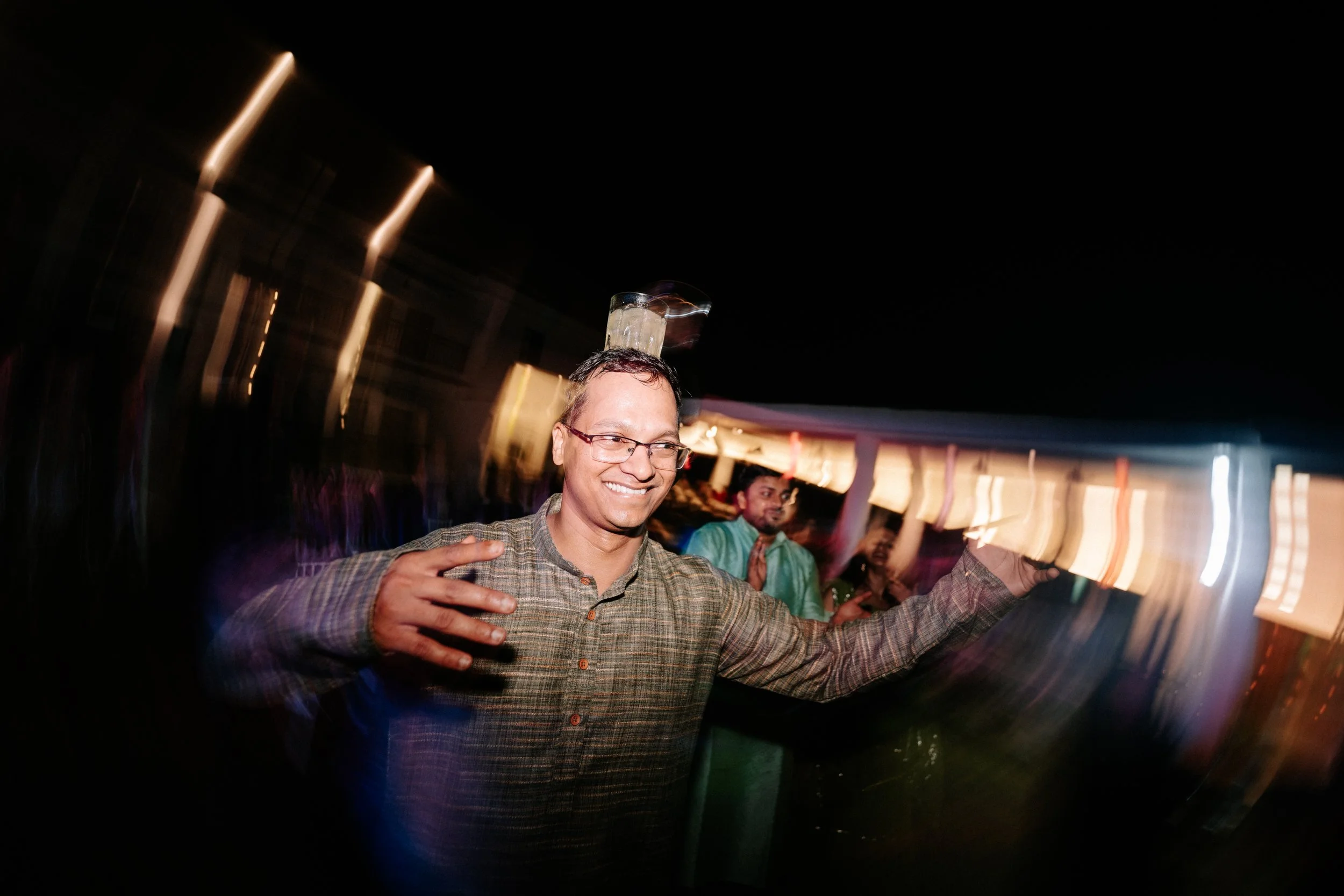 A man smiling with glasses and a brown plaid shirt balancing a glass on his head at a nighttime outdoor event, with colorful blurred lights and other people in the background.
