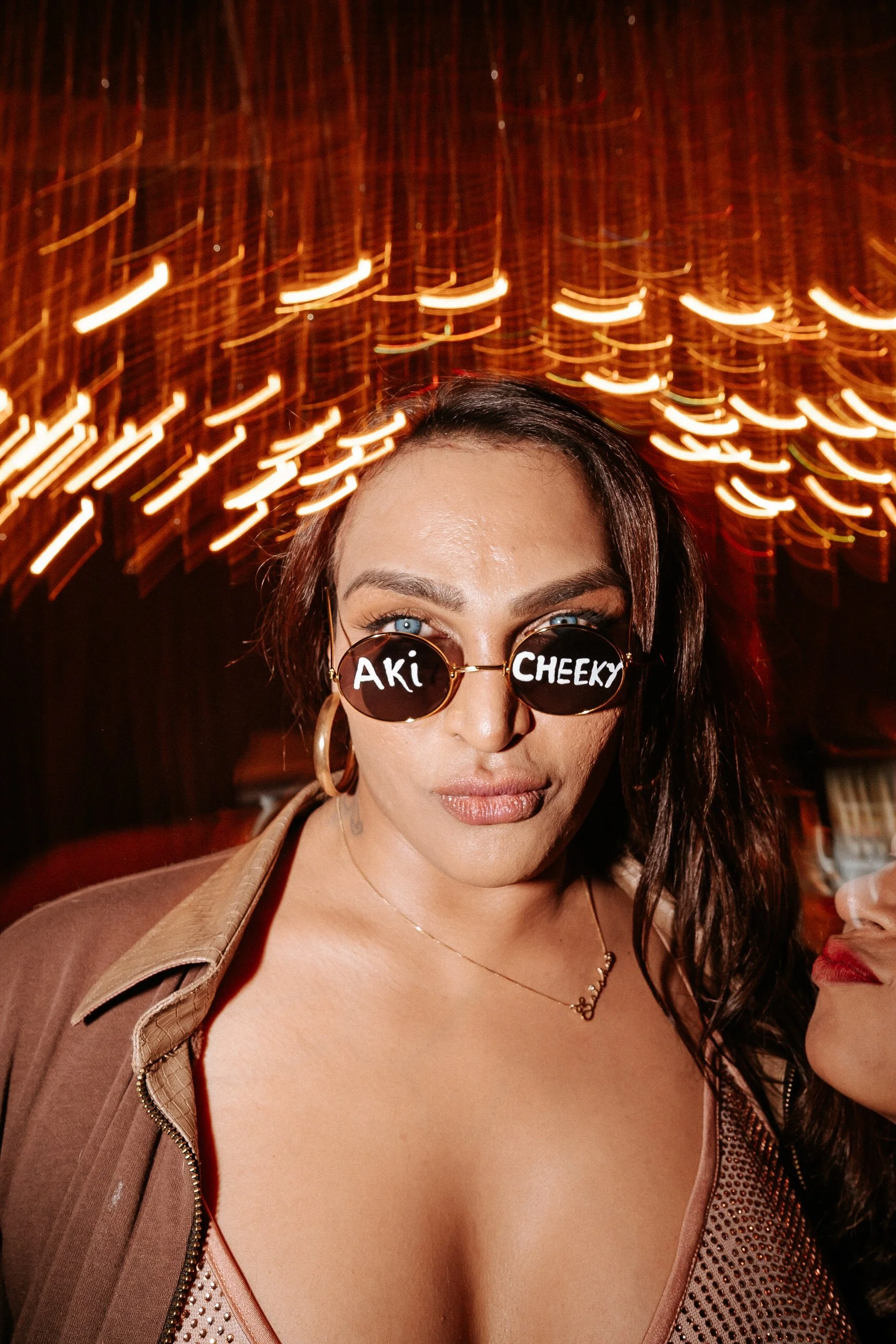 A woman with dark hair wearing sunglasses with words 'Aki' and 'Cheeky' on the lenses, in a dimly lit setting with blurred orange lights above.