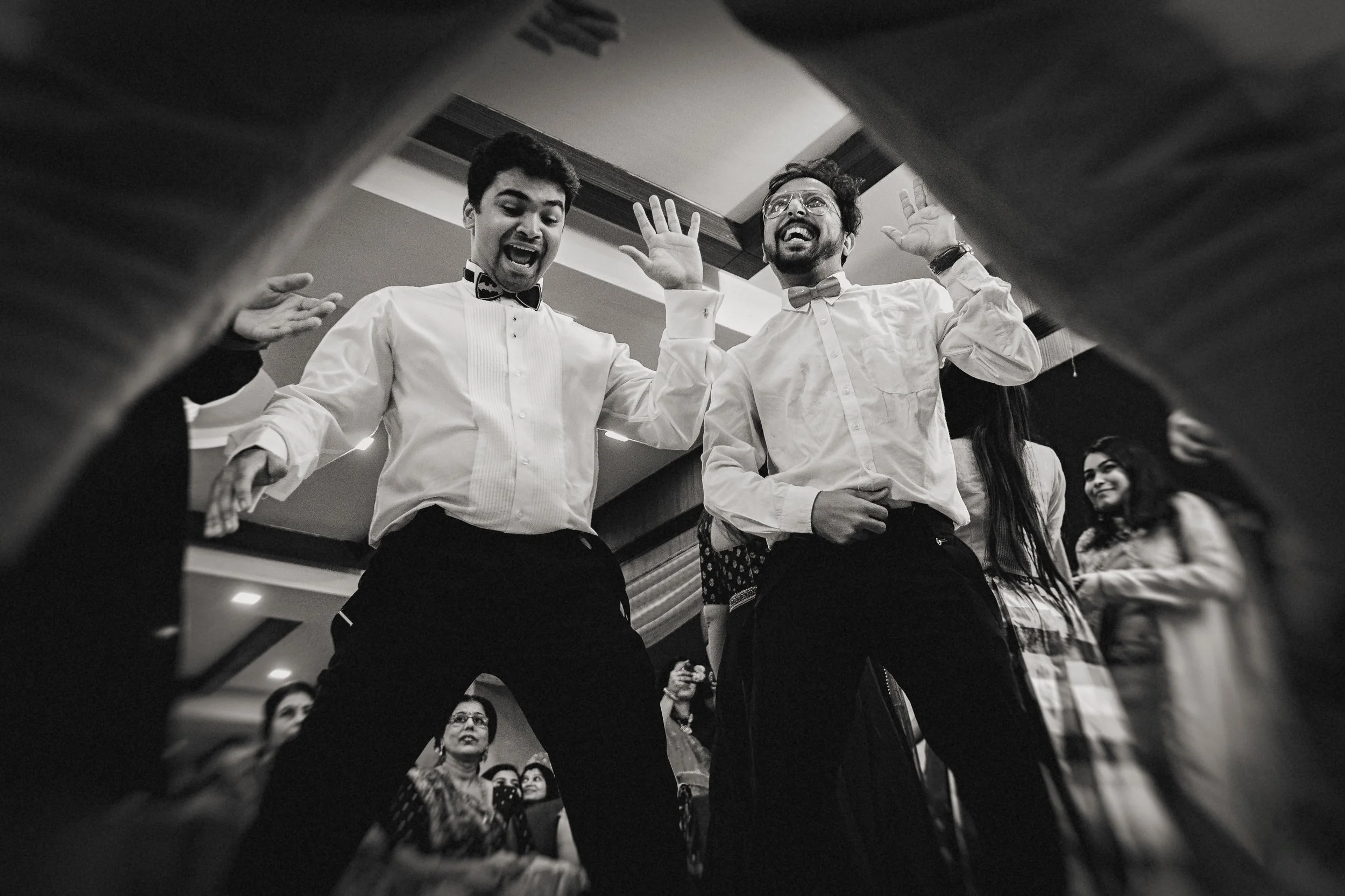 Two men in formal attire dancing and celebrating at a party, surrounded by other guests, captured from below through a camera lens.