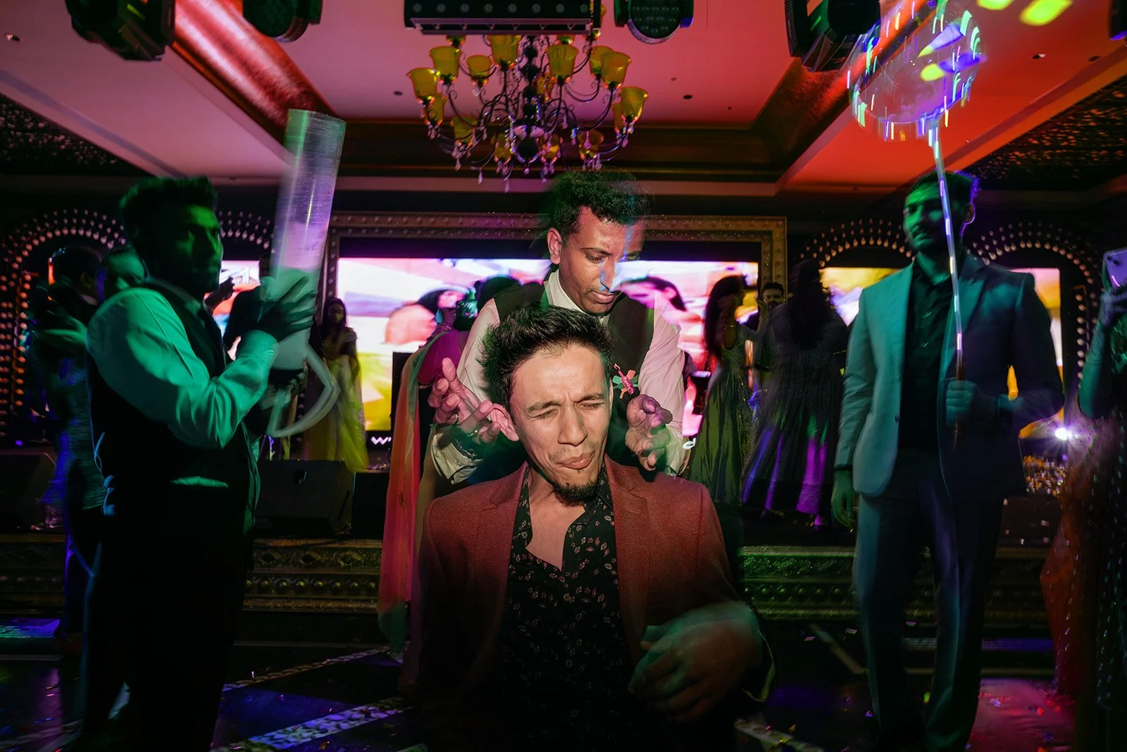 People dancing and celebrating at a party with colorful lights and a chandelier, with a man getting a massage or facial at the center.