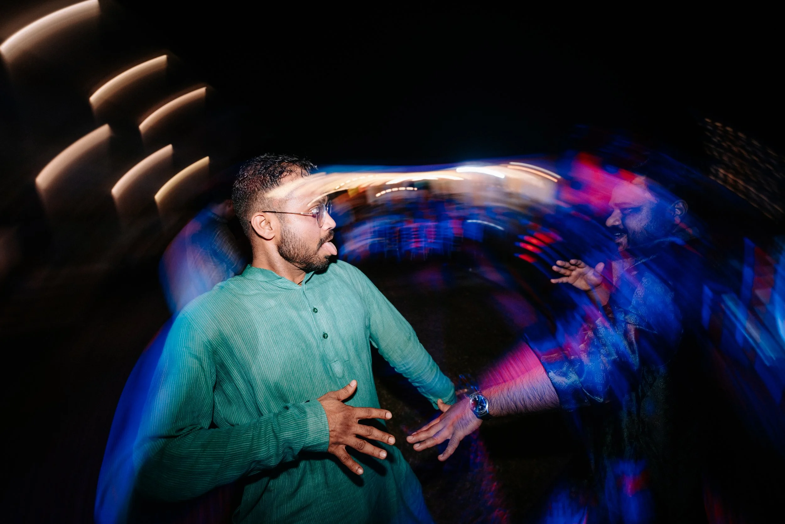 Two men at a party or nightclub, one wearing glasses and a green shirt, the other in a dark shirt, engaging in a conversation with motion-blurred colorful lights in the background.