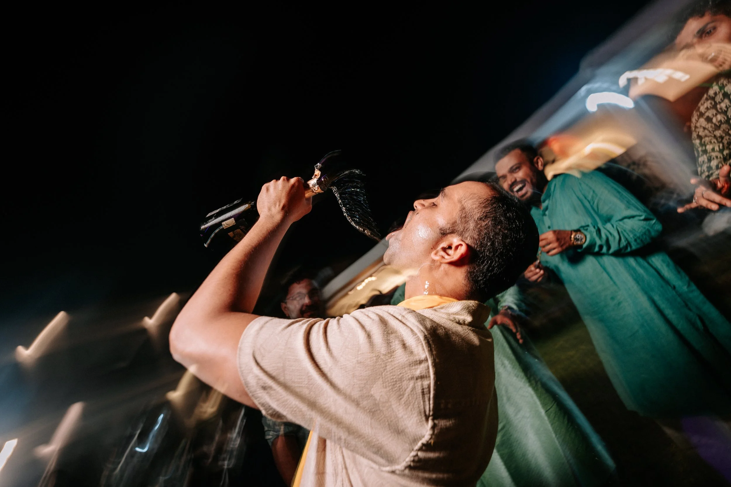 A man is celebrating with a bottle of champagne, pouring it into his mouth, while others surrounding him cheer and smile at a party or celebration at night.