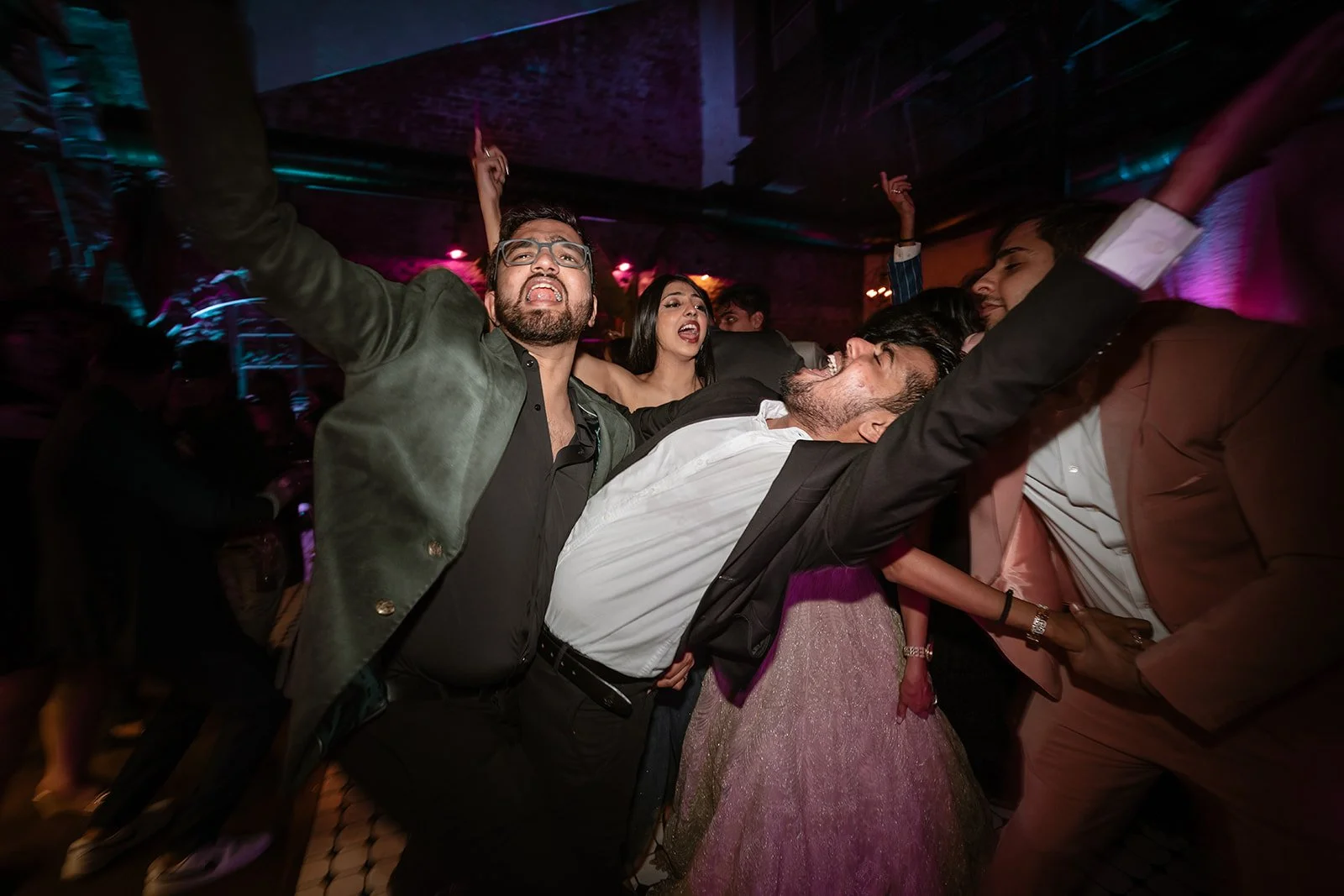 People dancing and celebrating at a lively party or nightclub, with a colorful purple and pink lighting atmosphere.