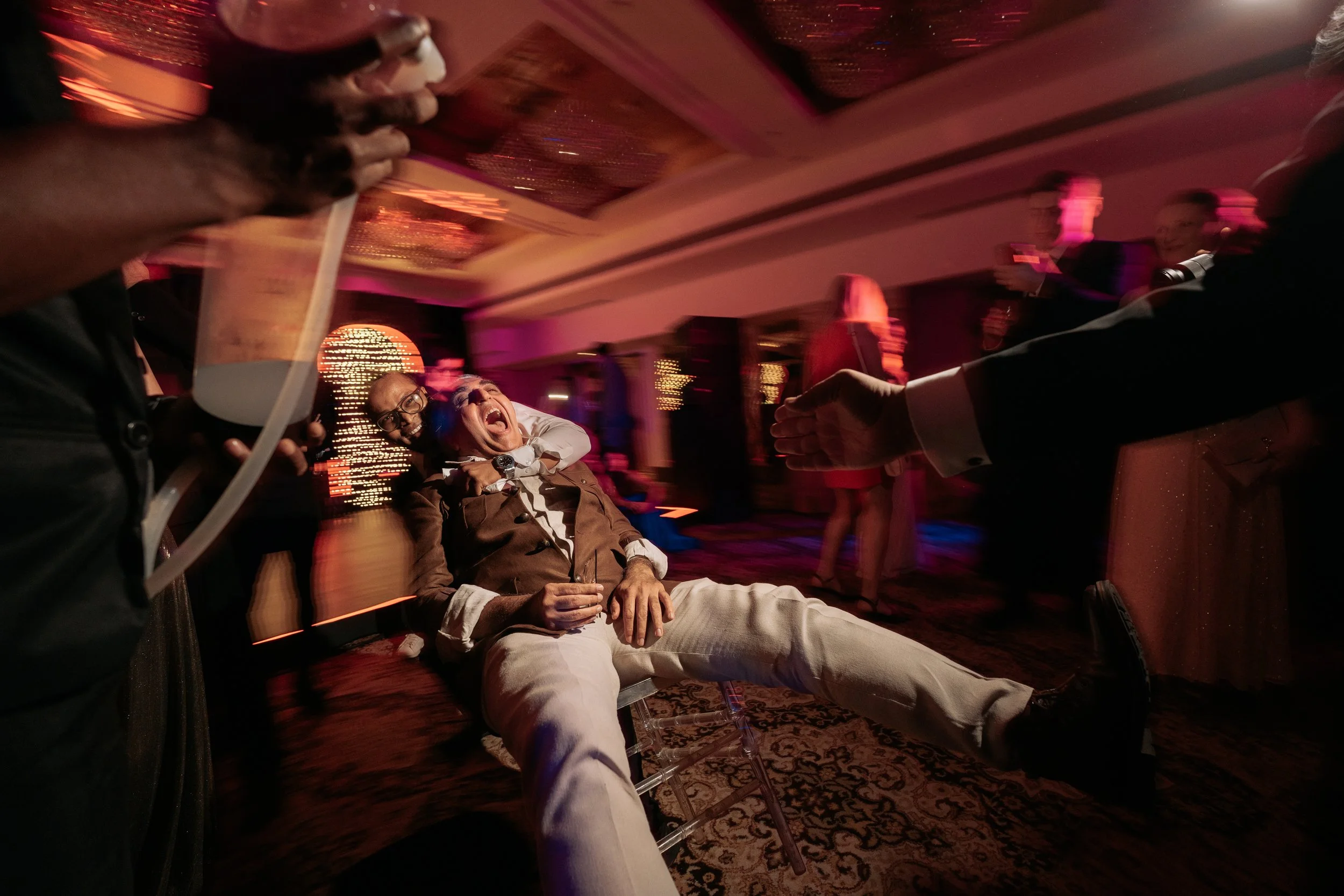 A man sitting in a chair laughing and shouting with his arms up, while a woman with red hair stands in the background at a lively party or celebration, with colorful lighting and people dancing in the background.