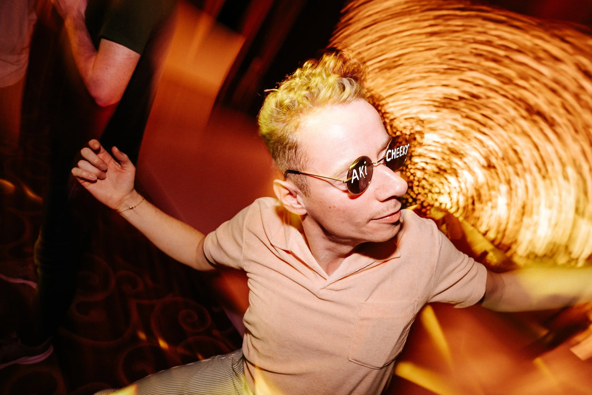 A person with blond curly hair and sunglasses that read "AKL CHECK" is dancing or posing at a lively event, with warm lighting and a patterned floor in the background.