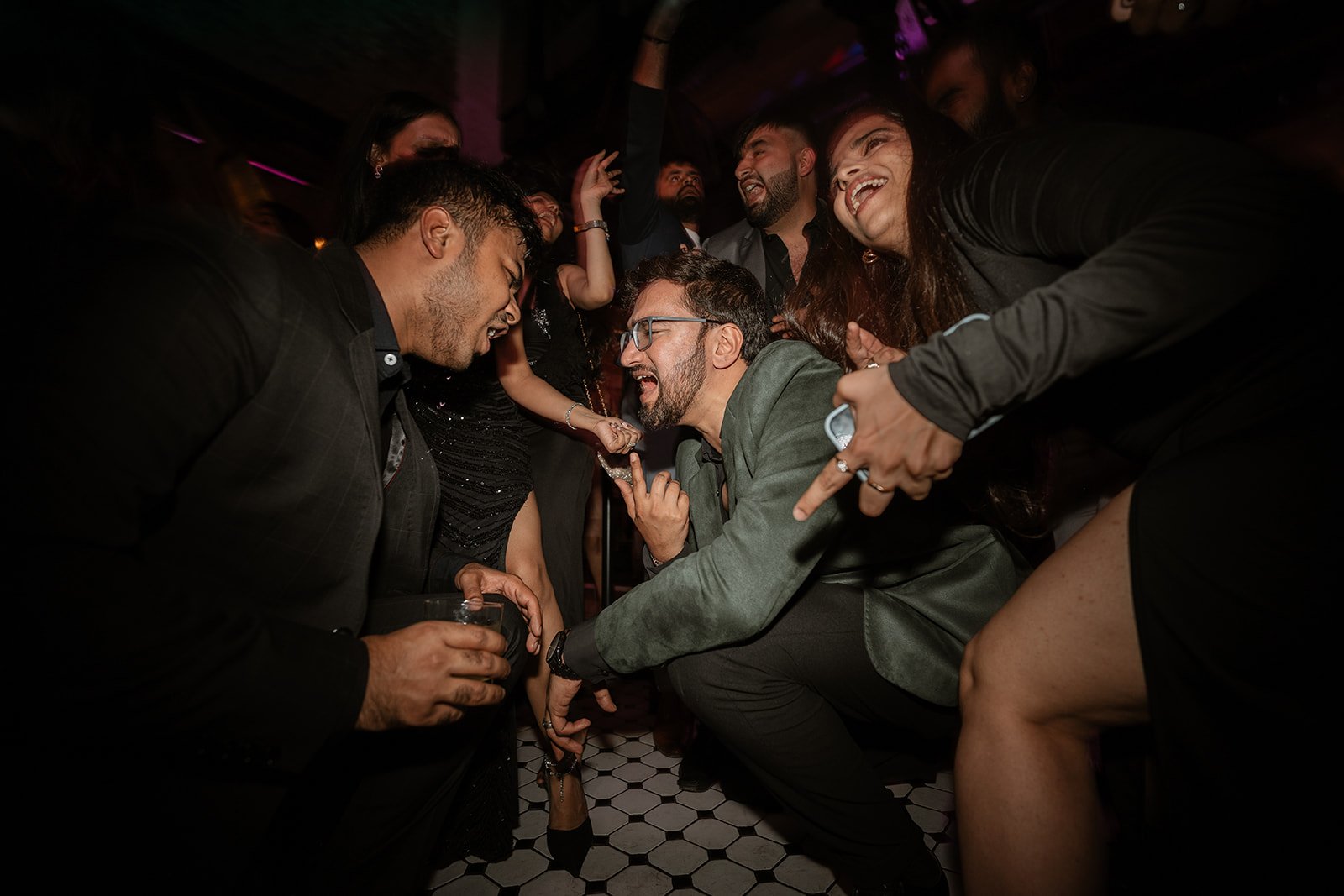 A group of people dancing and laughing at a party with dim lighting, drinks in hand, and a black-and-white tiled floor.