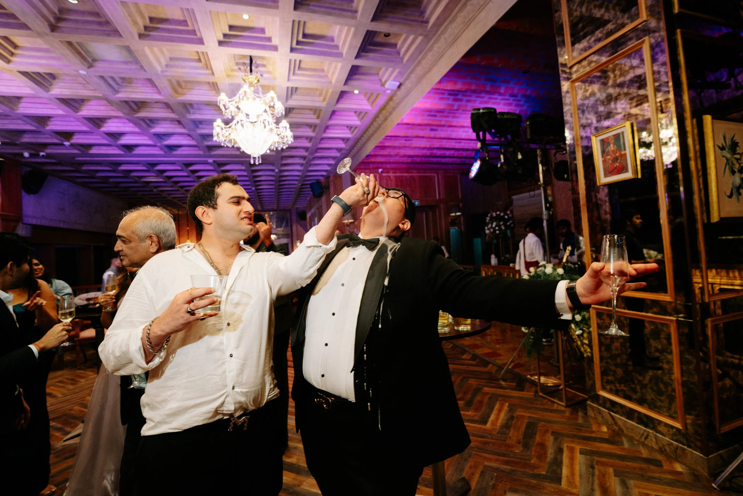 People celebrating at a party, with two men in the foreground, one feeding the other with a glass, in a decorated indoor setting with a chandelier and framed artwork.