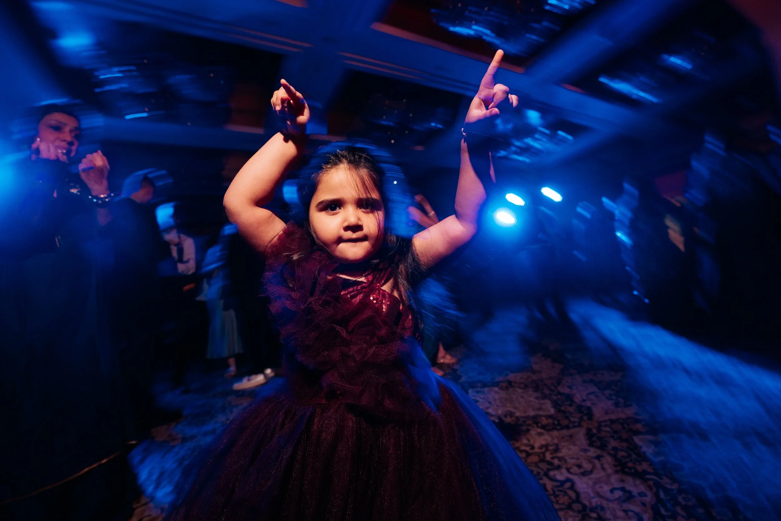 A young girl in a dark purple dress dancing at a party with blue and purple lights, with other people dancing in the background.
