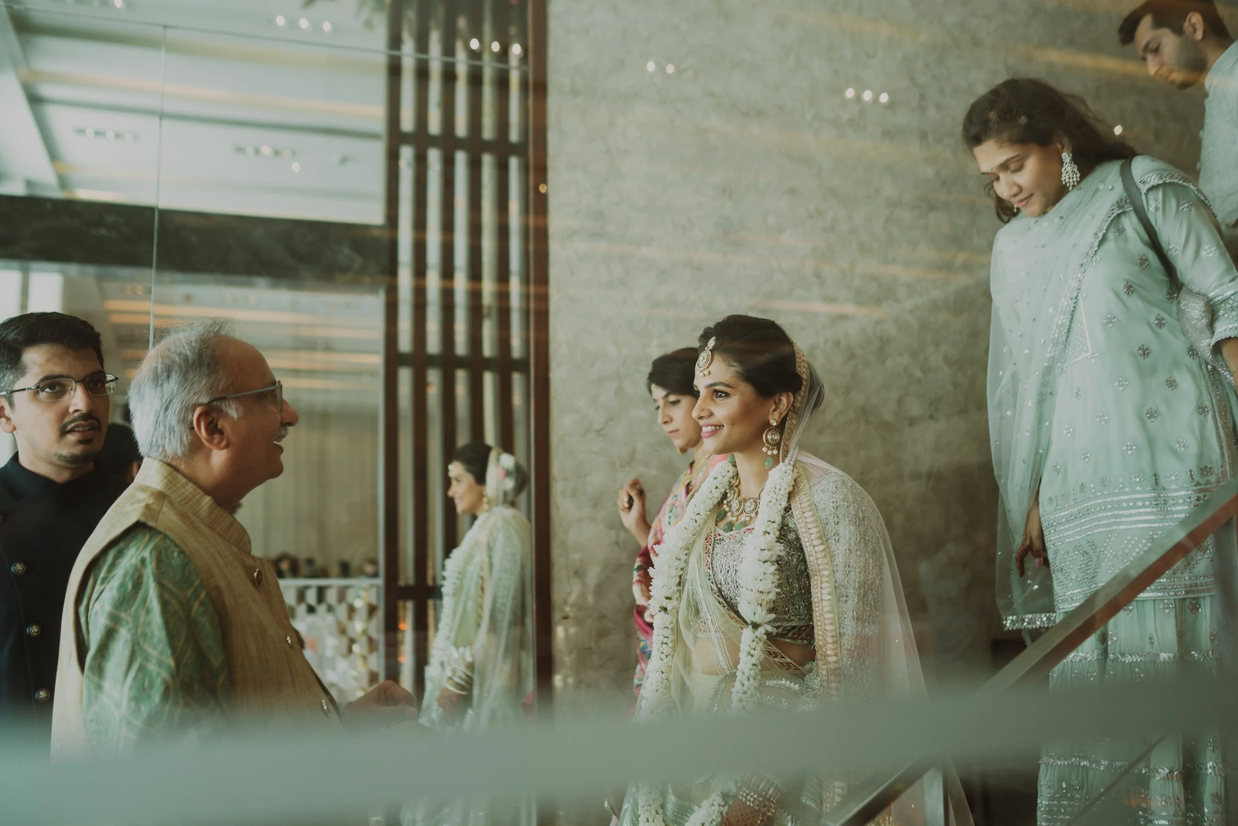 An Indian bride wearing traditional jewelry and attire, smiling and talking to an elderly man, with women in traditional dresses and a man in the background at a wedding or event.