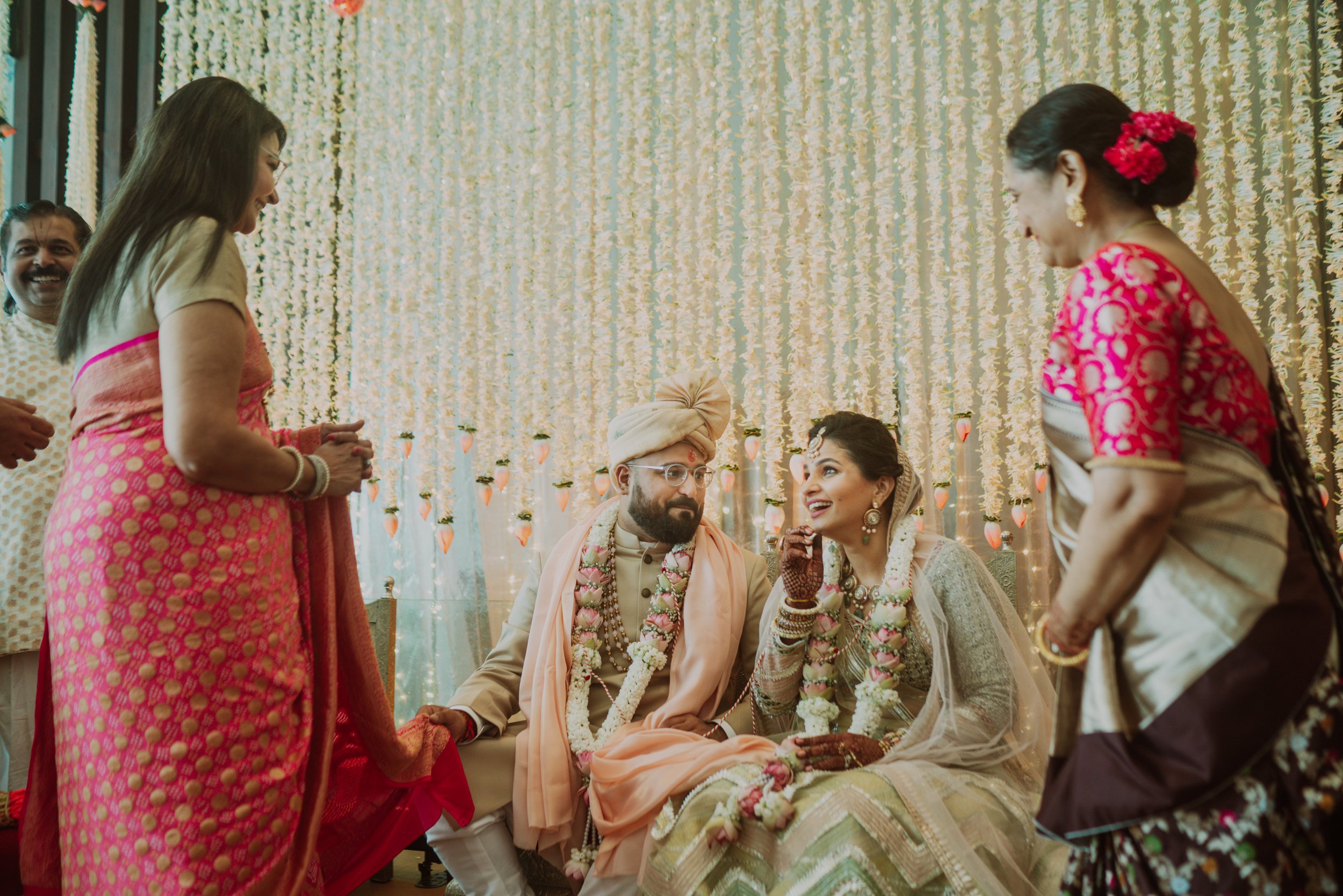 Indian wedding ceremony with the bride and groom sitting together, surrounded by family members, in front of a decorated backdrop with strings of flowers and hanging lanterns.