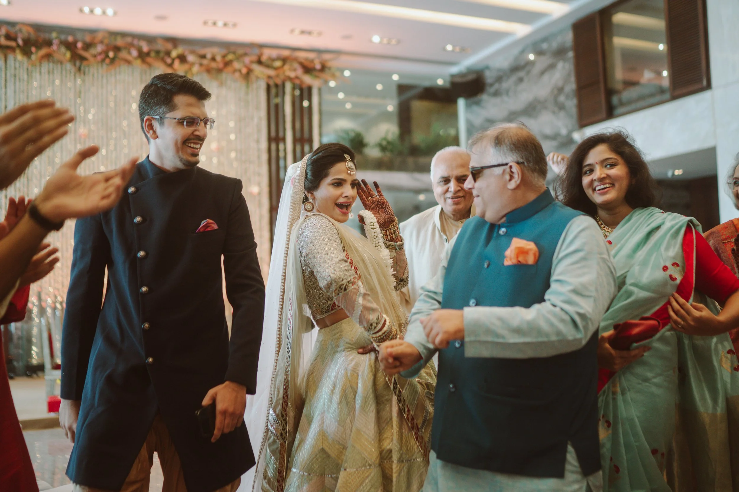 People celebrating an Indian wedding, with a bride in traditional attire and family members smiling and clapping in a decorated indoor venue.