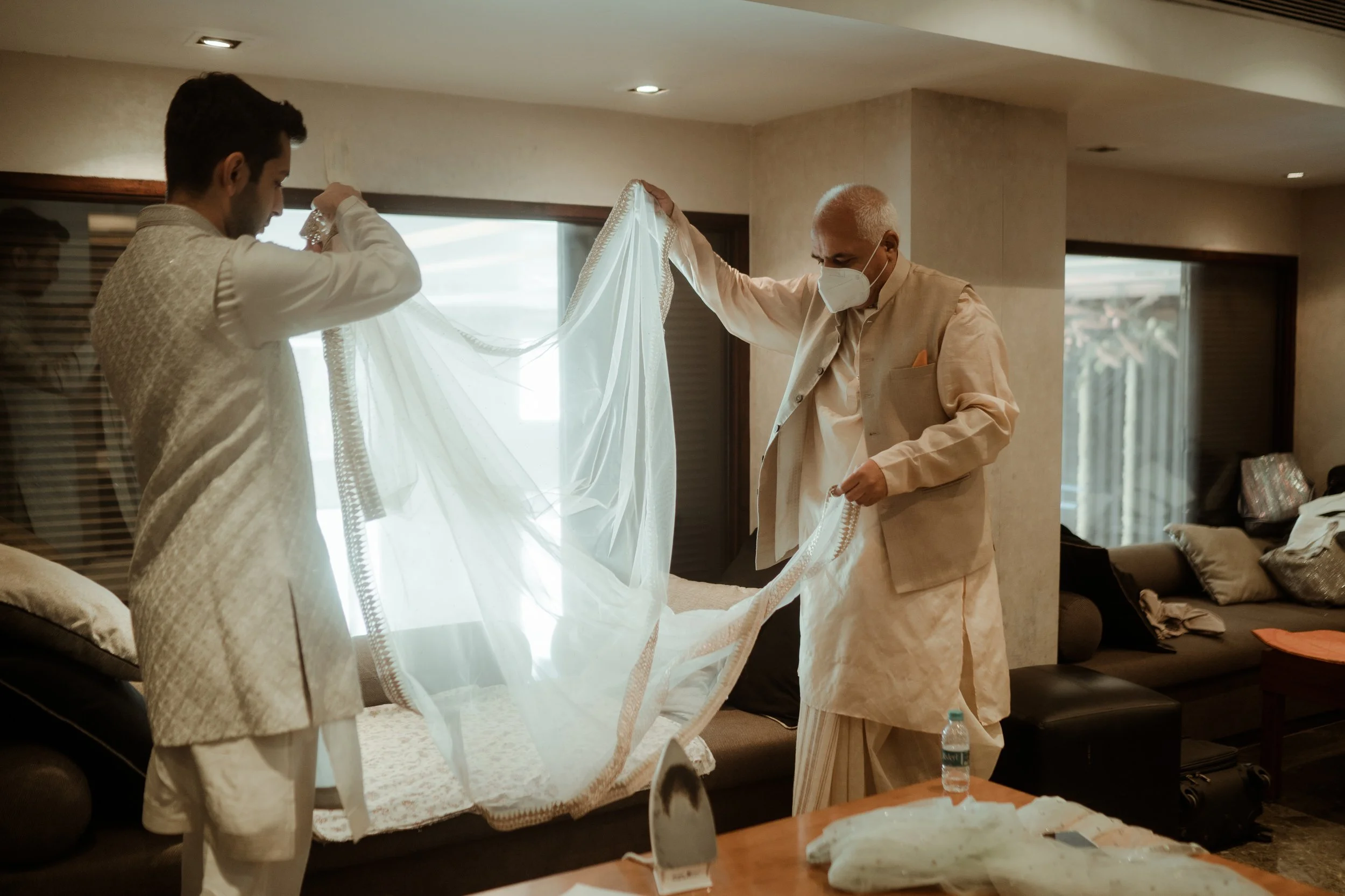 Two men in traditional Indian attire placing a white wedding veil on a sofa, with one wearing a face mask, in a living room with sunlight coming through windows.