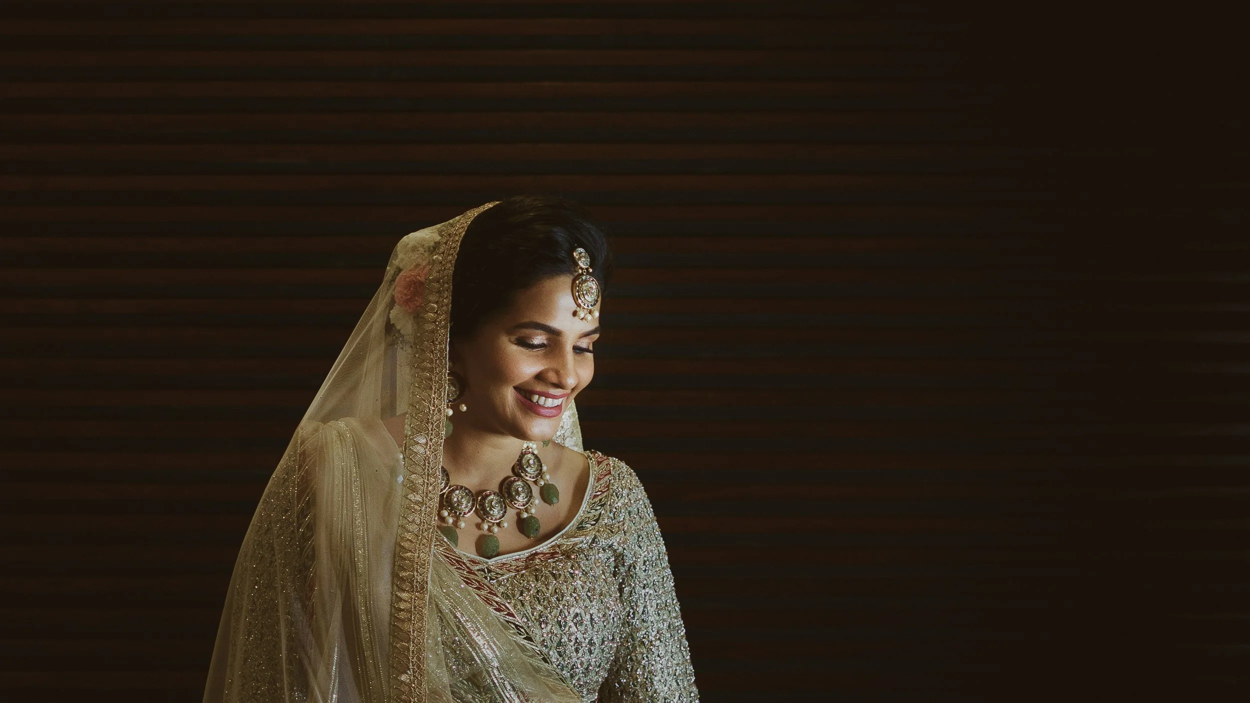 Elegant woman dressed in traditional Indian bridal attire, smiling with eyes closed, wearing ornate jewelry including a necklace, earrings, and headpiece, with a decorative veil over her head, against a dark wooden background.