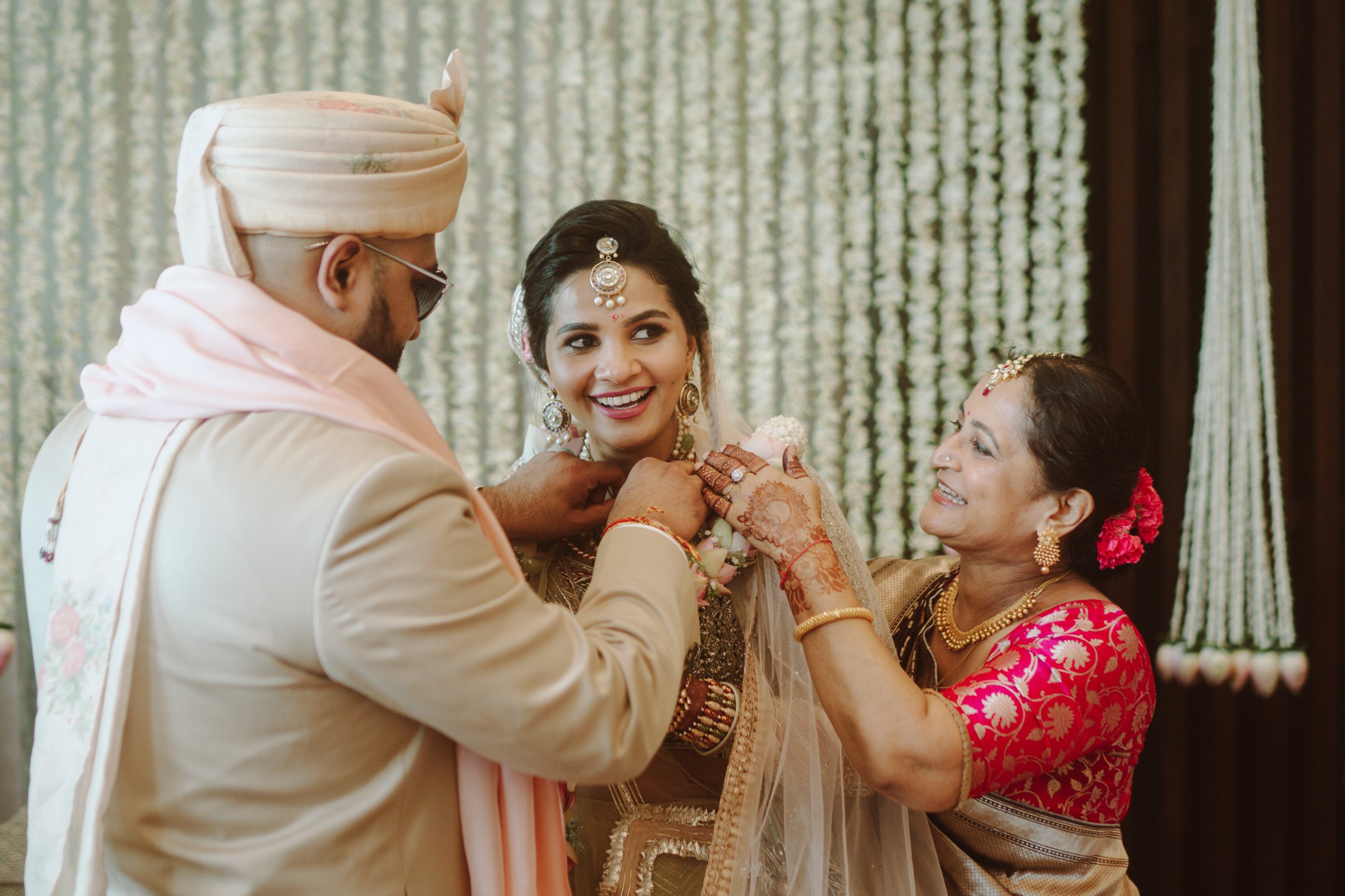 A bride in traditional Indian wedding attire being adorned with jewelry by her mother and a man in traditional Indian attire at a wedding ceremony.