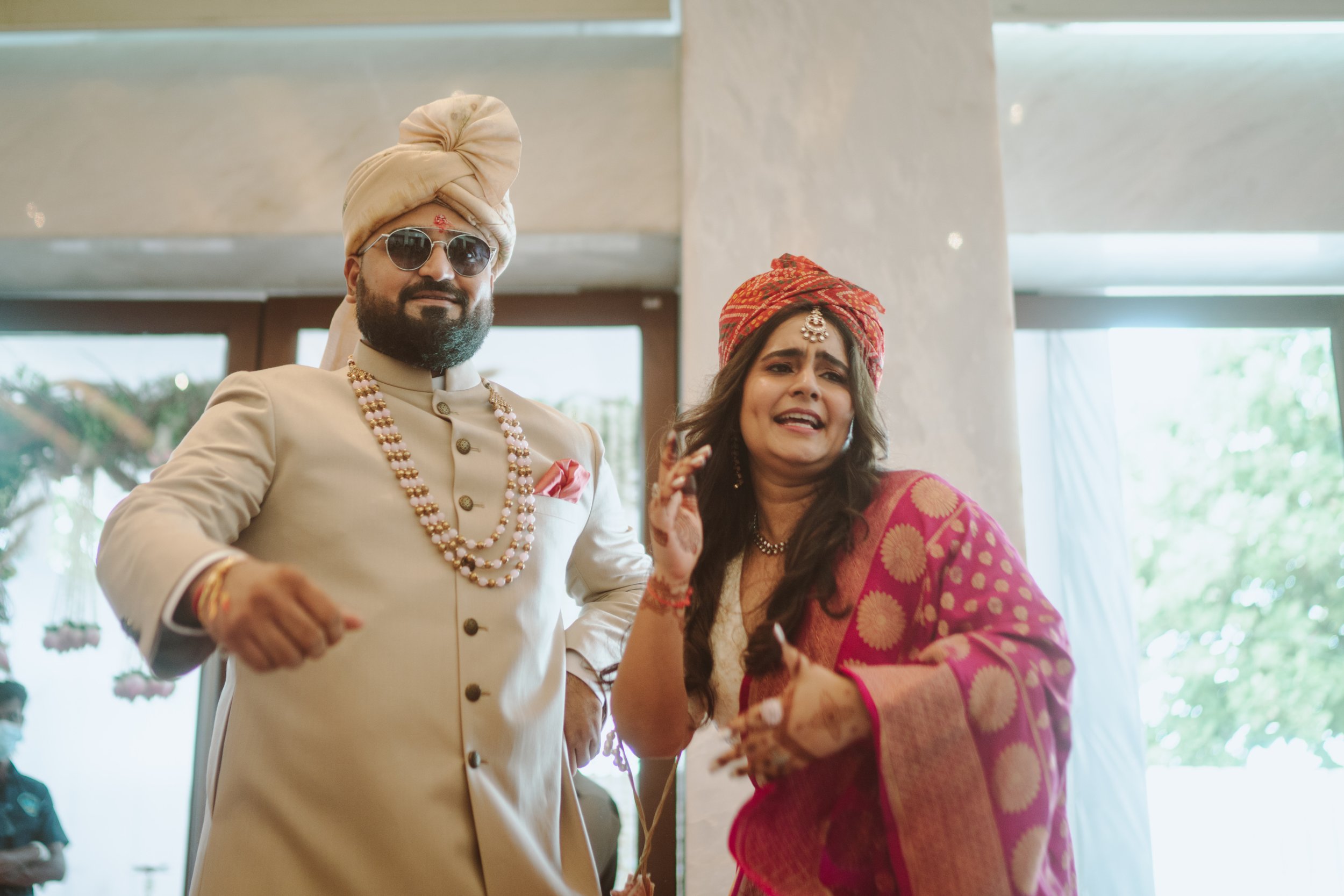 A man dressed in traditional Indian wedding attire with sunglasses and a turban, standing next to a woman in colorful traditional Indian clothing and headscarf, both smiling and trying to dance at a wedding celebration indoors.