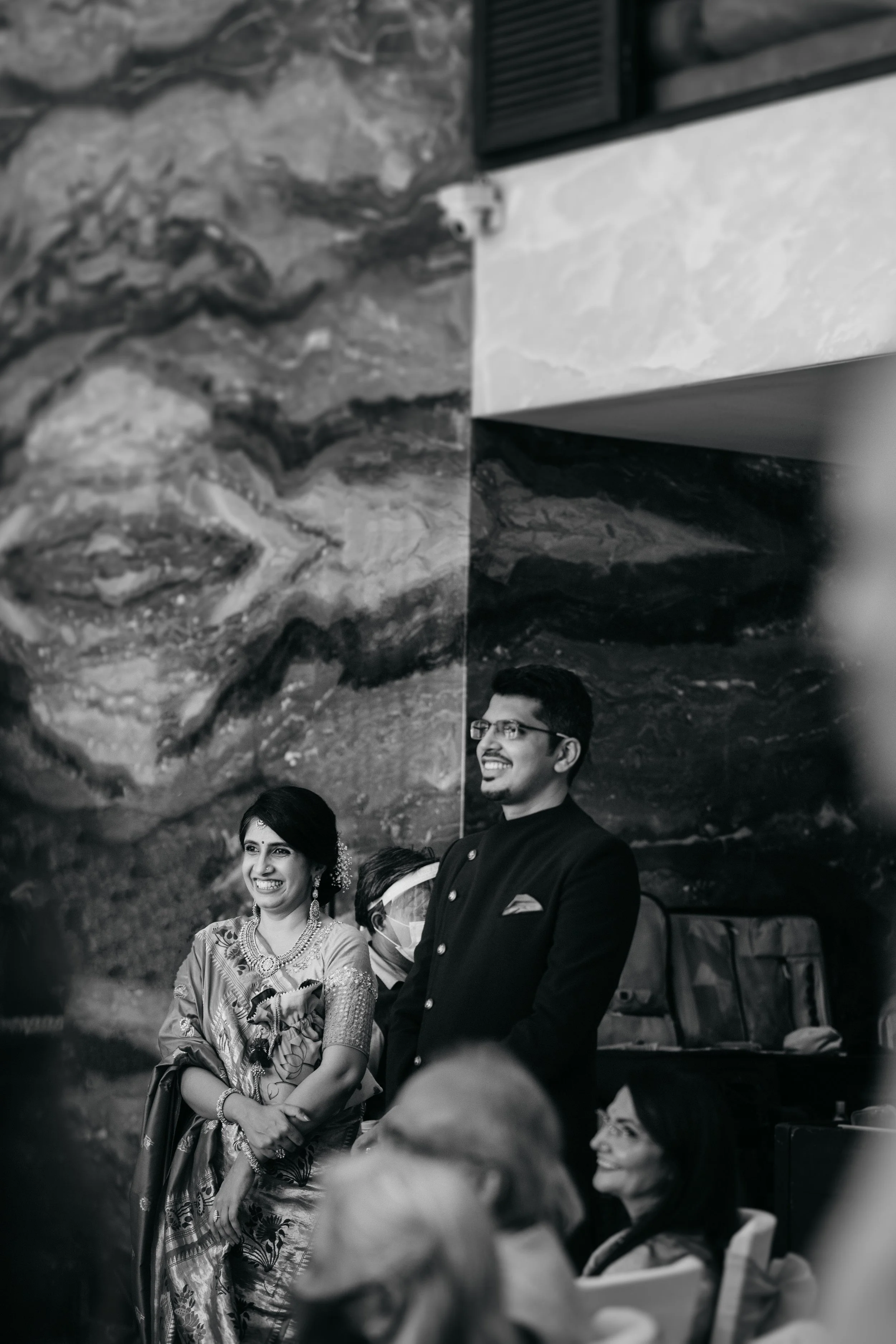 A black and white photograph of a celebration, featuring a smiling woman dressed in traditional Indian attire, and a smiling man in formal attire standing beside her, with other people seated and smiling in the background.