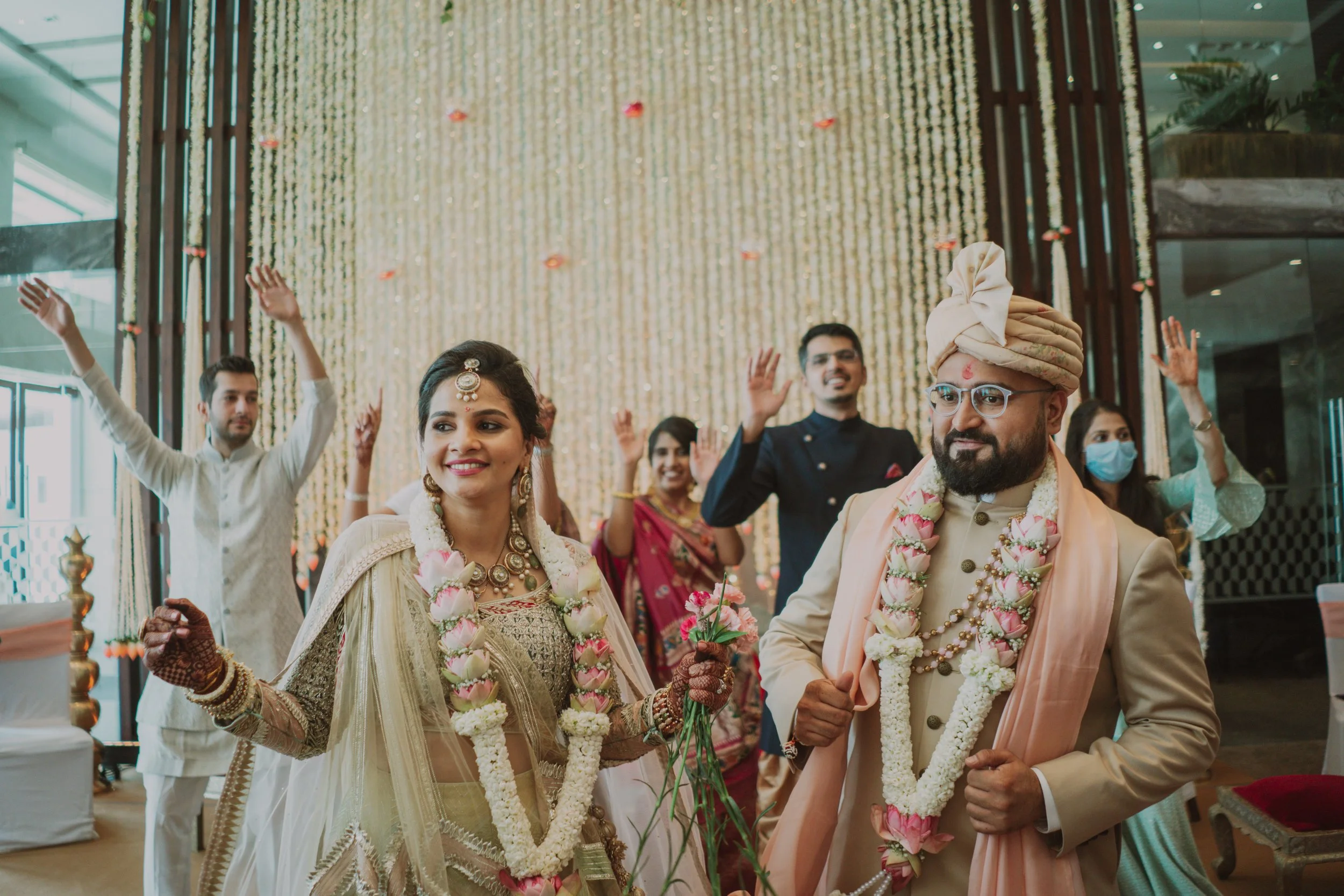 Indian wedding celebration with bride and groom wearing traditional attire, floral garlands, and jewelry, surrounded by family and friends in festive mood.