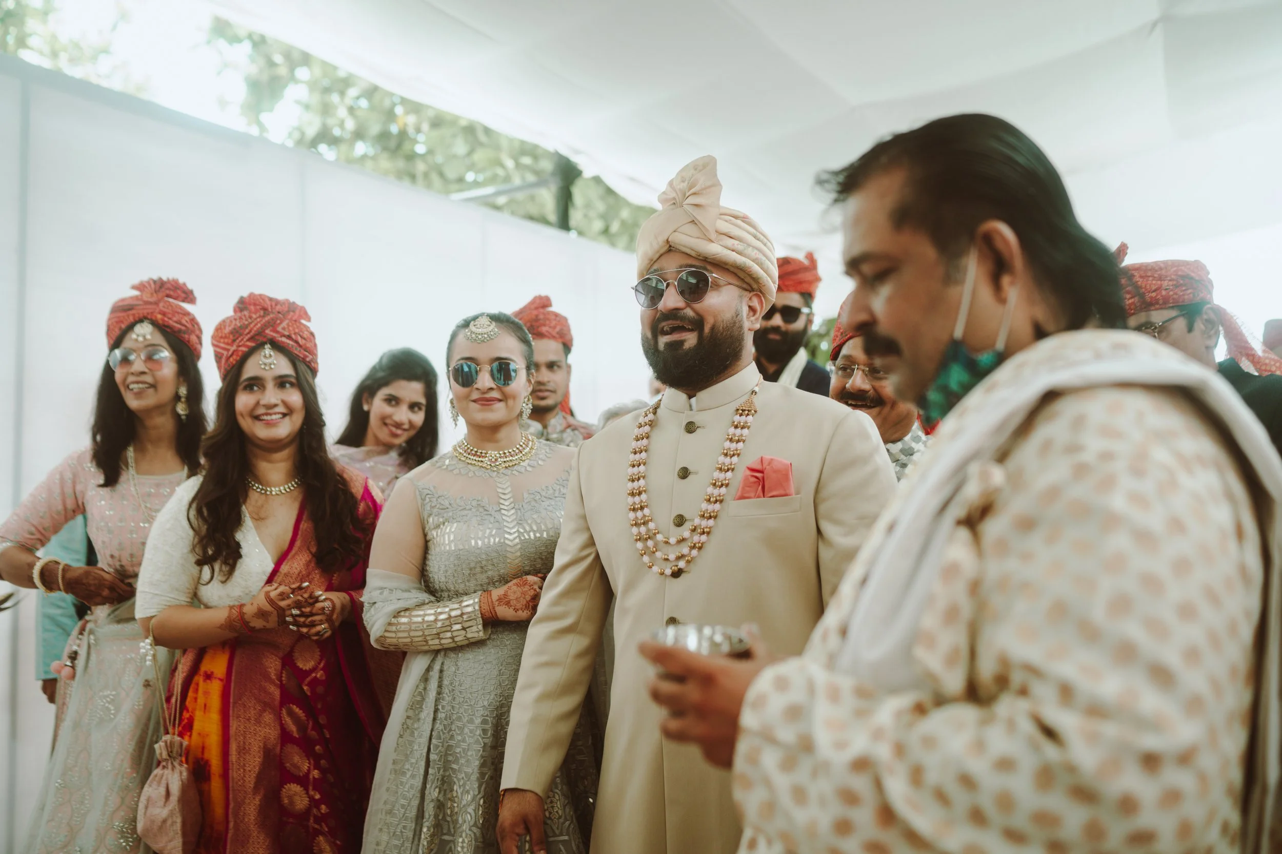 Indian wedding ceremony with men and women dressed in traditional attire, some wearing turbans and jewelry, gathering under a white canopy.