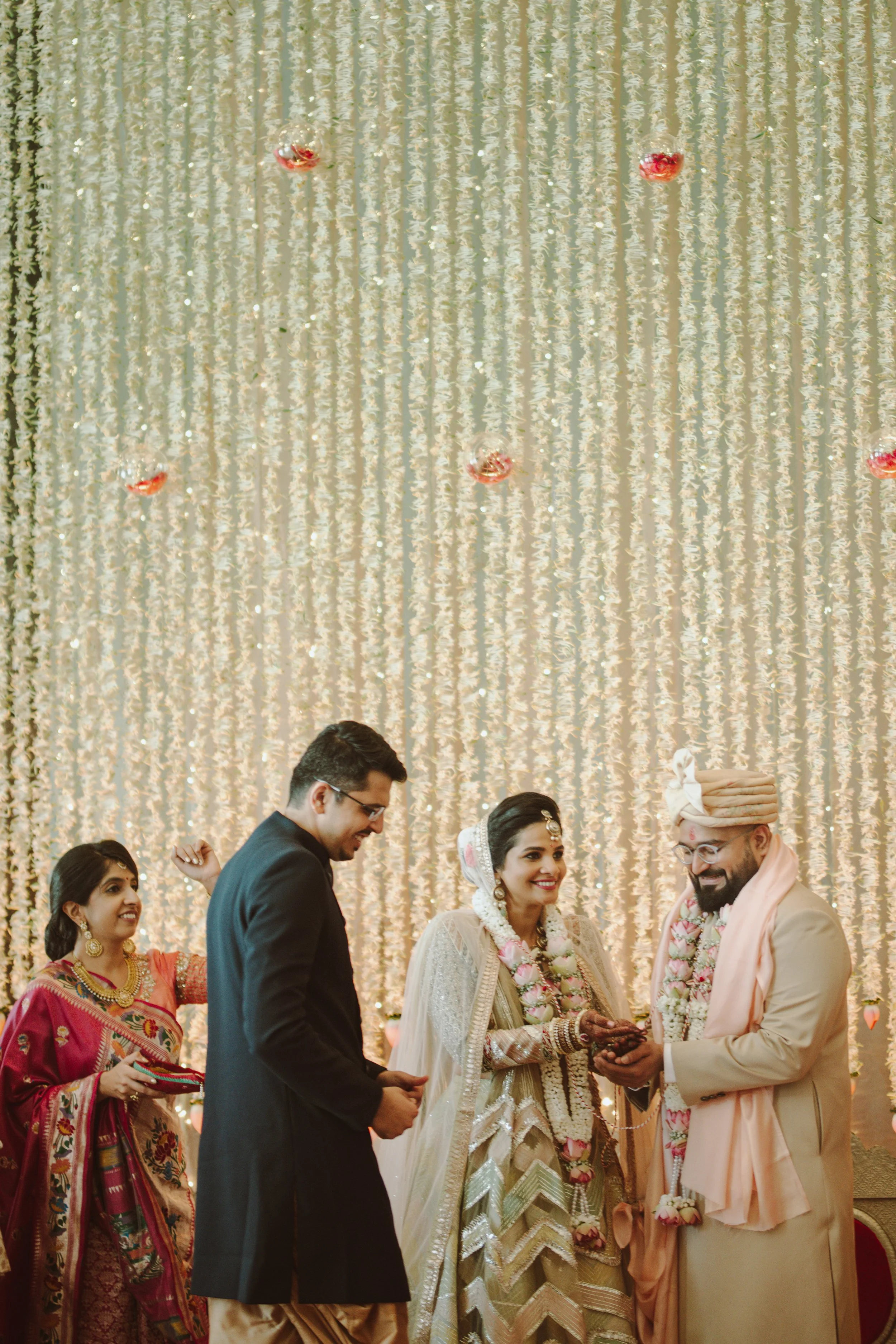 Indian bride and groom exchanging rings during wedding ceremony with friends and decorative backdrop.