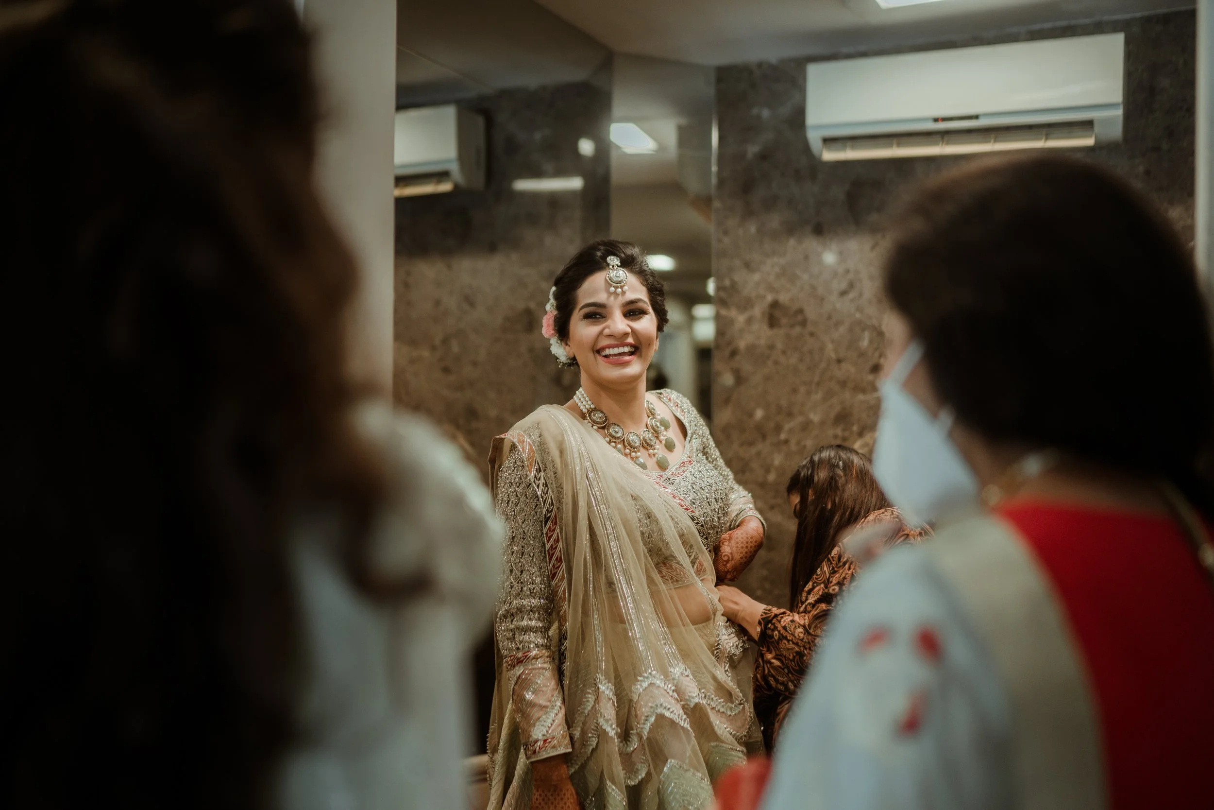 A smiling woman dressed in traditional Indian wedding attire, wearing jewelry and floral hair accessories, is surrounded by other women, one of whom is wearing a mask, in an indoor setting with stone walls and air conditioning units.
