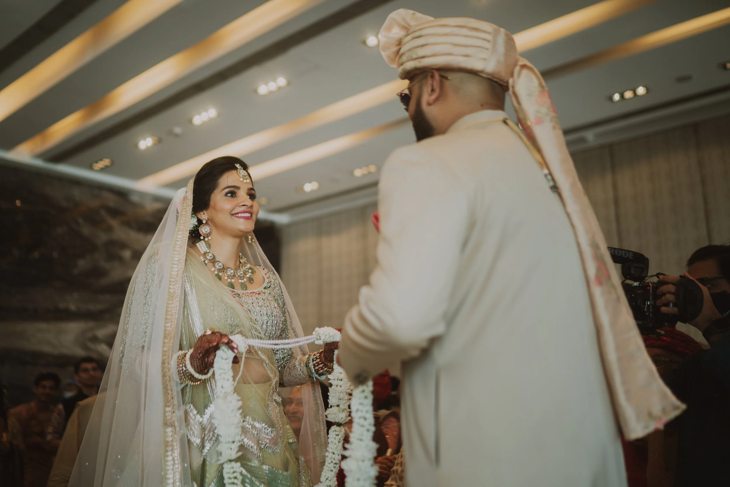 Bride and groom exchanging garlands during a traditional wedding ceremony, with the bride smiling and wearing elaborate jewelry and a veil, and the groom in a cream-colored outfit and turban.
