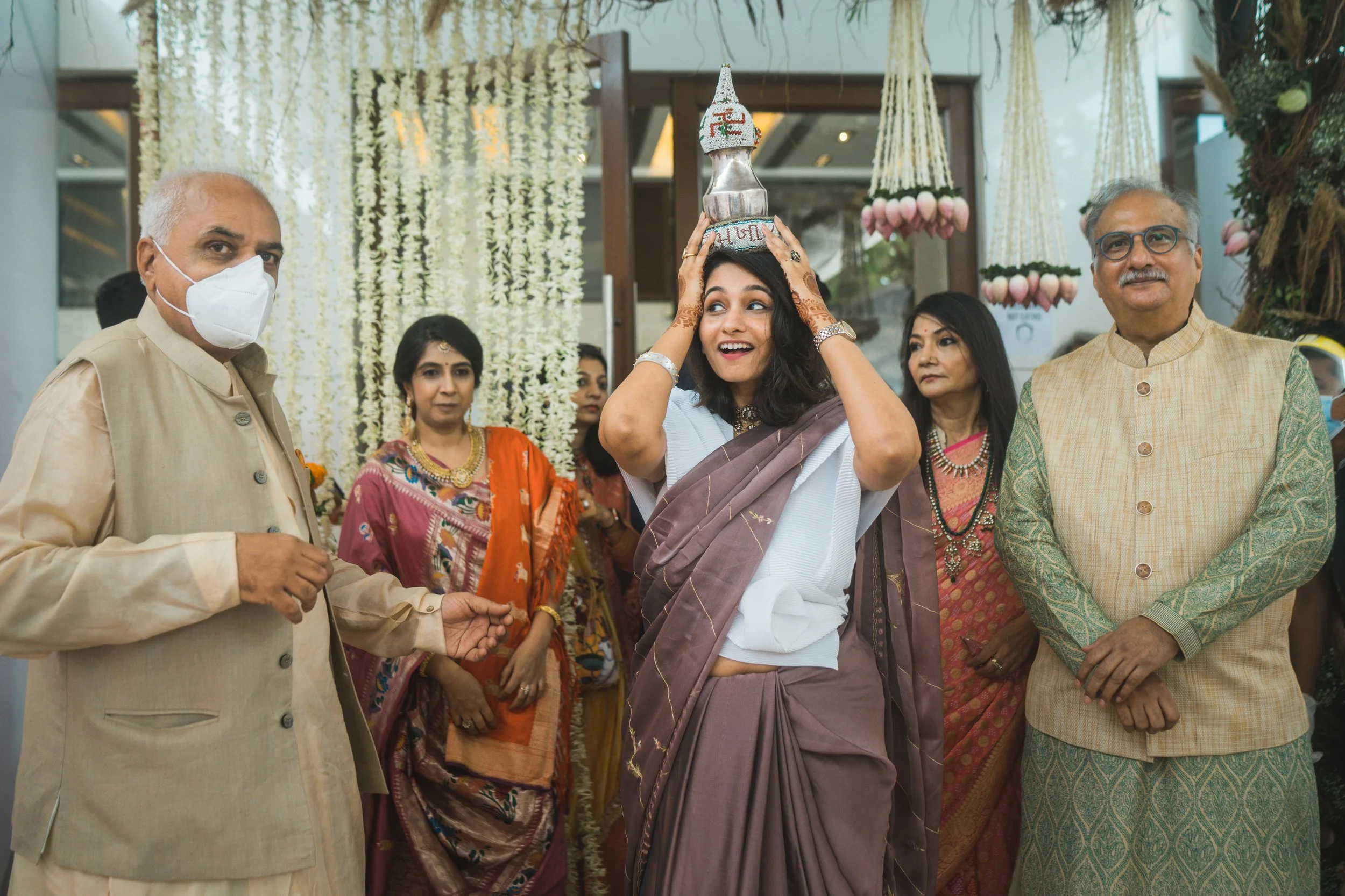A woman in traditional Indian attire is balancing a ceremonial object on her head, surrounded by men and women in traditional clothing at a festive event with floral decorations.