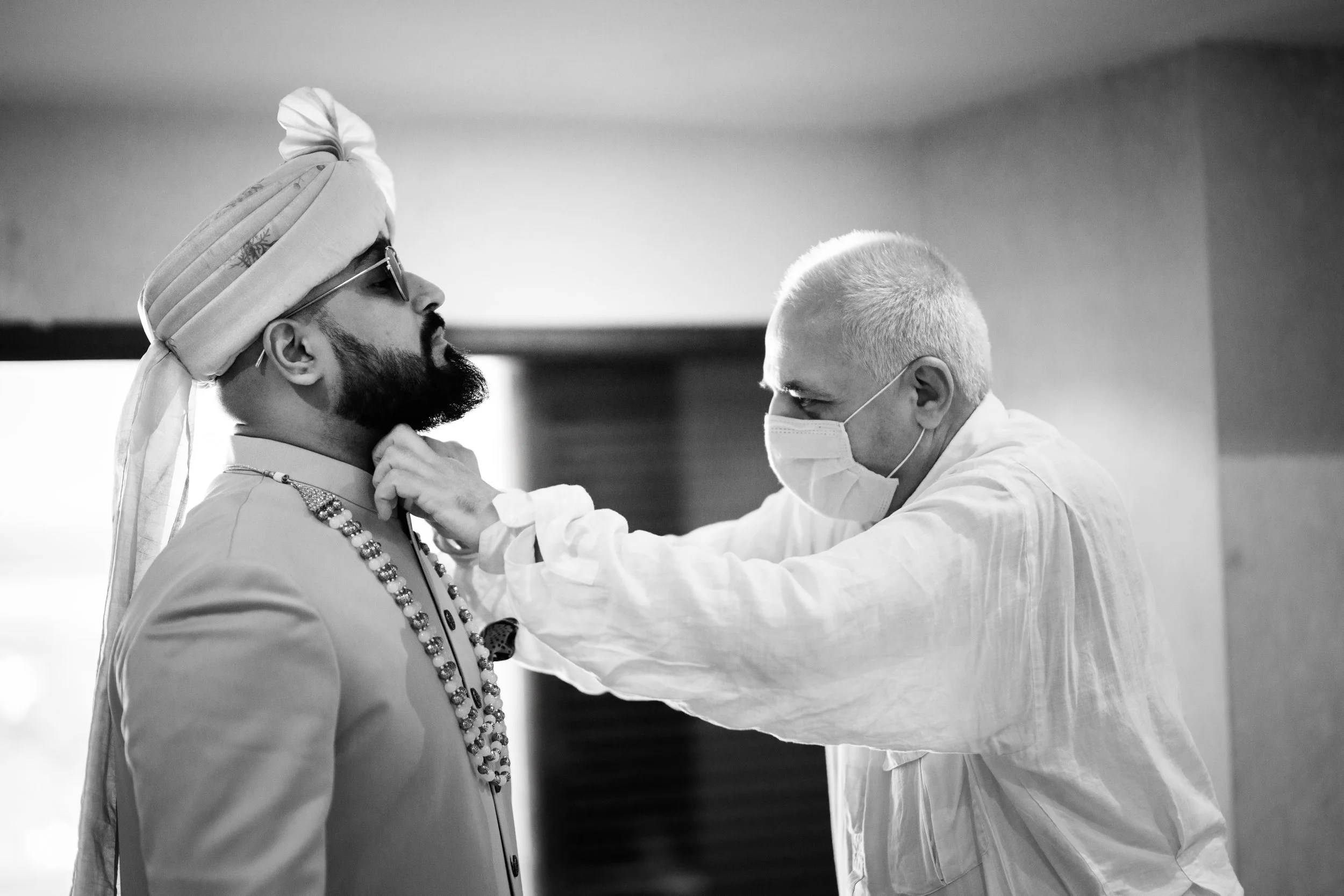 A man wearing traditional Indian attire and a turban is being assisted by a person wearing a medical mask, who appears to be adjusting or inspecting his clothing or jewelry.