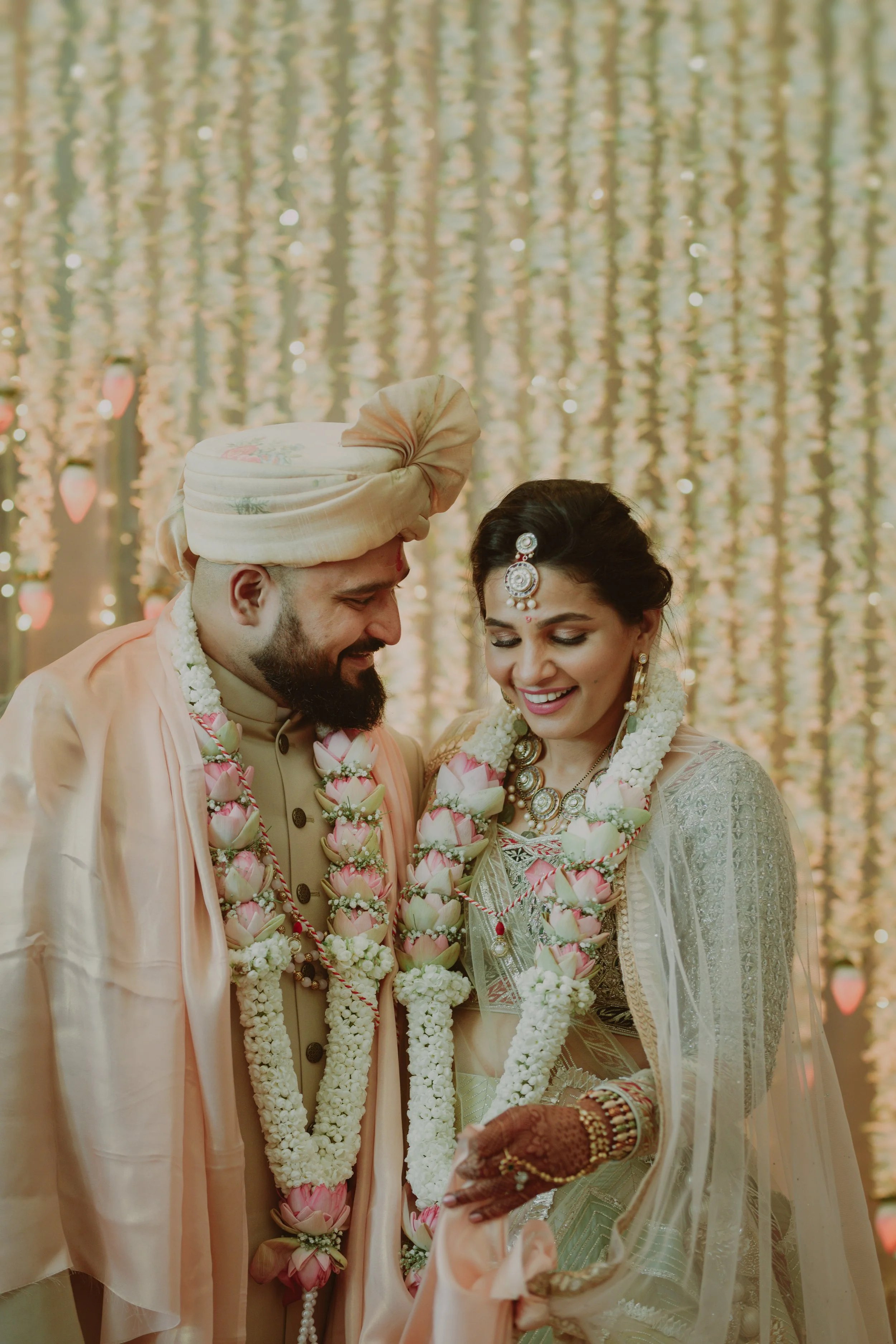 Indian wedding ceremony with bride and groom smiling in traditional attire, adorned with flower garlands and jewelry, standing in front of a decorated backdrop.
