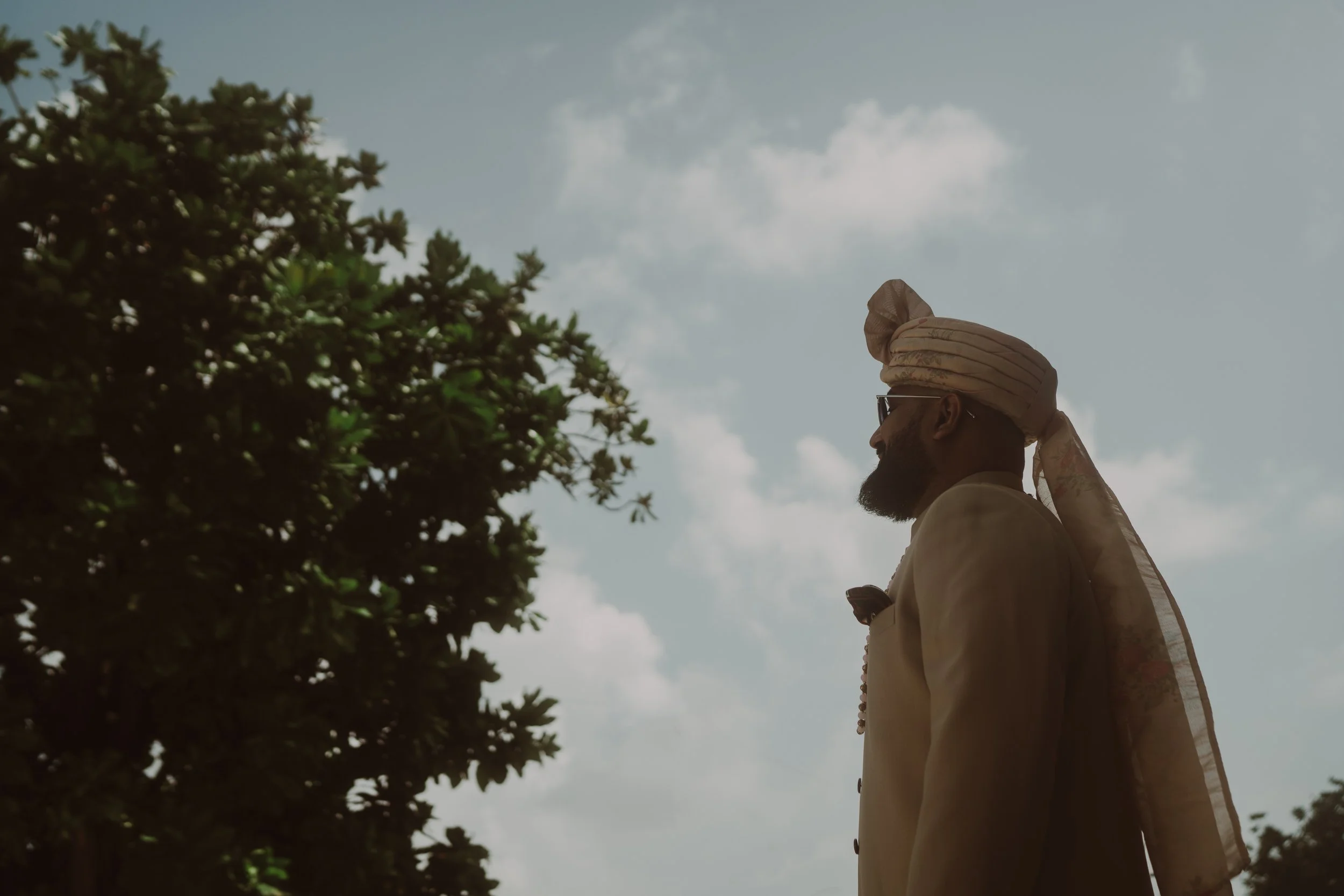 Profile of a man wearing a traditional turban and glasses, standing outdoors near a tree with a cloudy sky in the background.