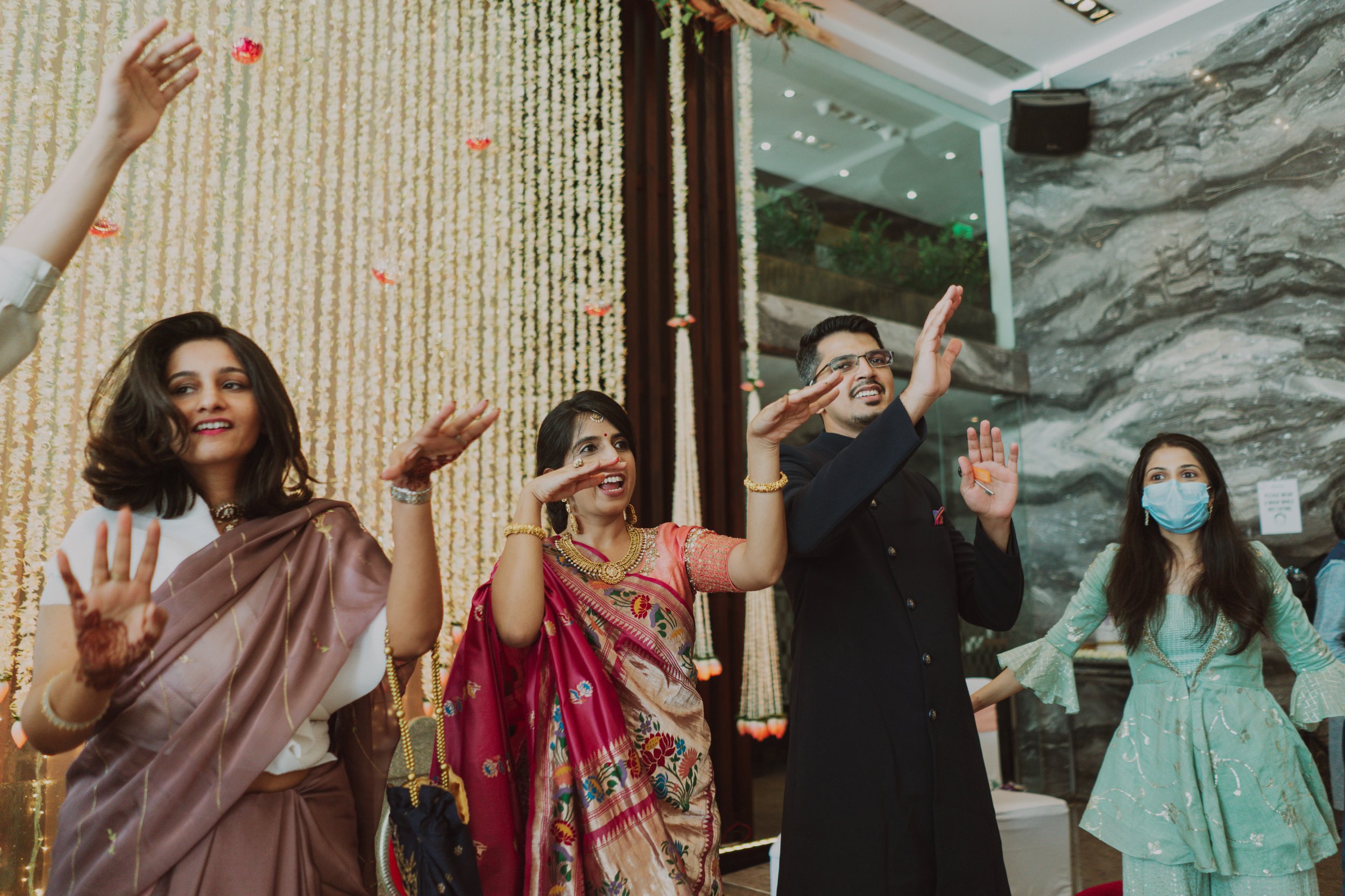 A group of people celebrating at a festive event, dressed in traditional Indian attire, some wearing masks, standing in front of a decorated backdrop.