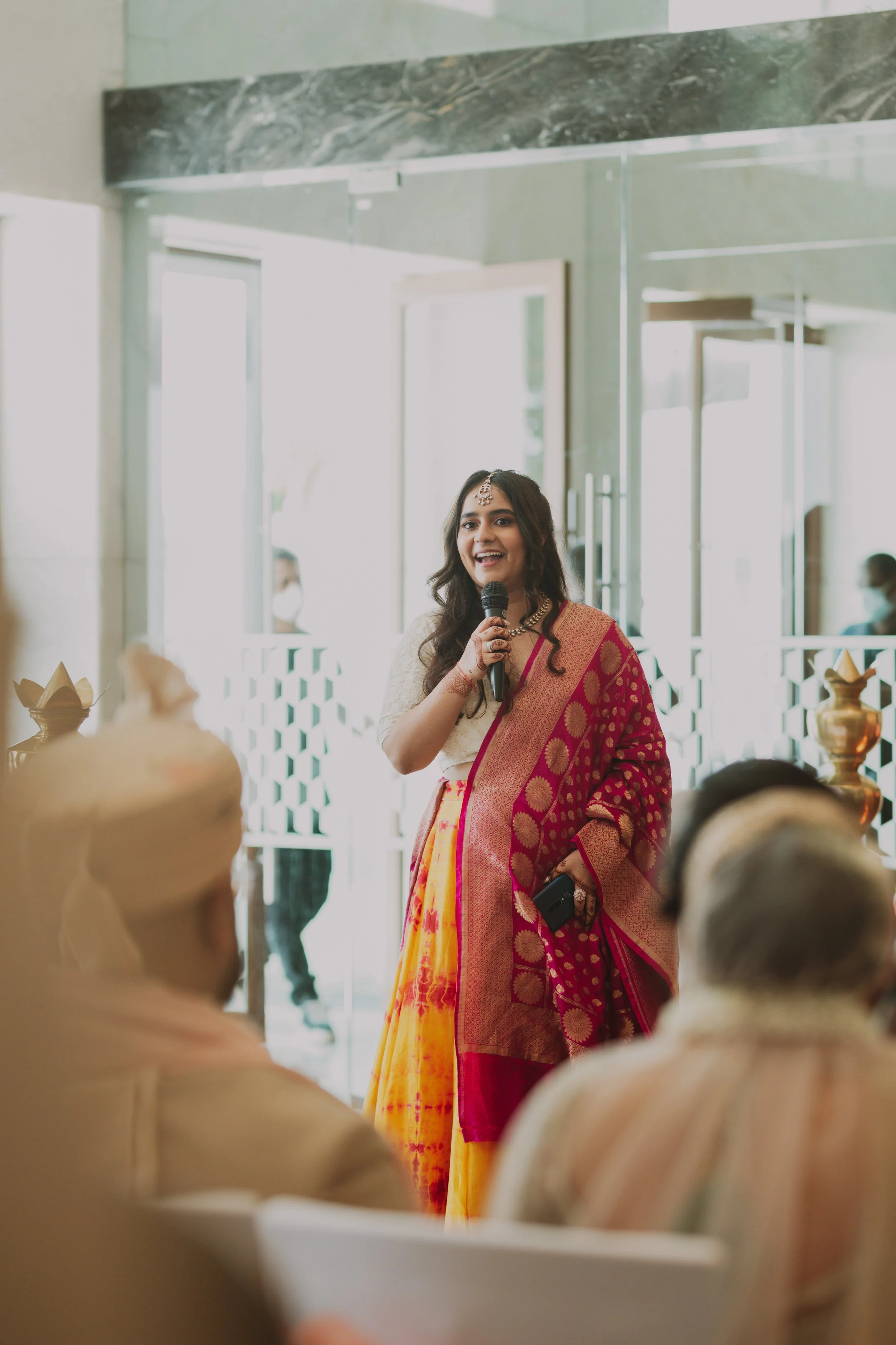 A woman in a traditional saree speaking into a microphone at an indoor event, with elderly men sitting in the foreground.
